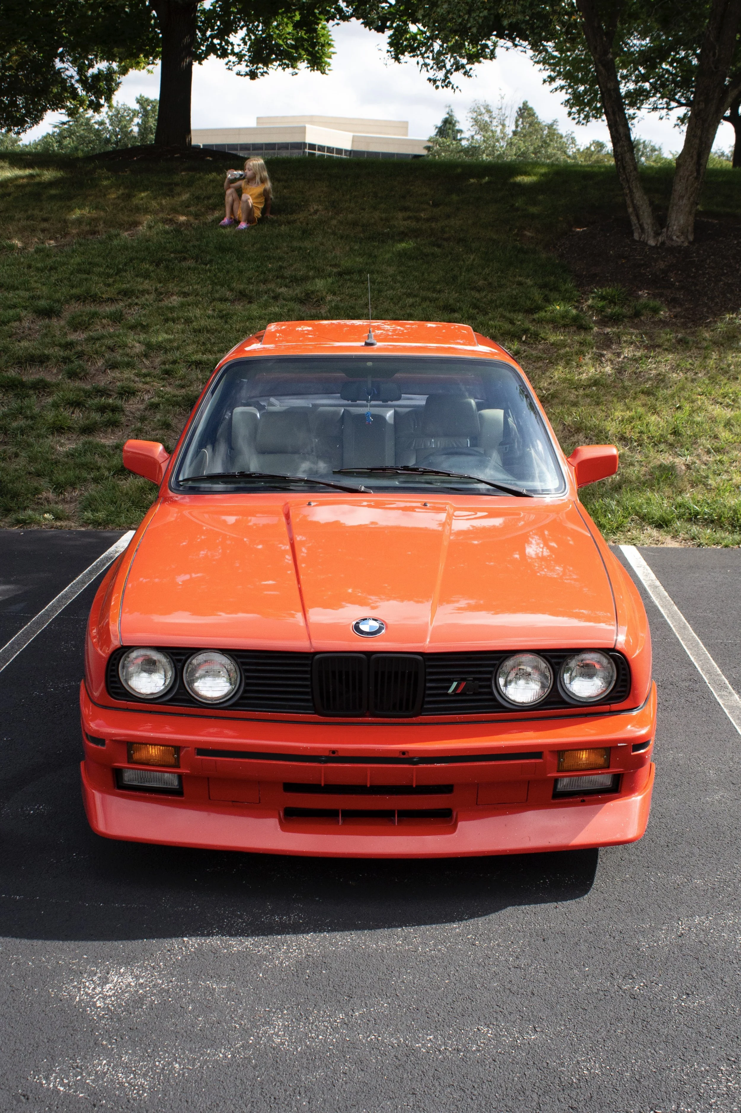 Front view of a bright orange vintage BMW car parked in a parking lot with white lines. In the background, a girl in a yellow dress is sitting on a grassy hill, drinking from a water bottle under trees, with a modern building visible behind her.
