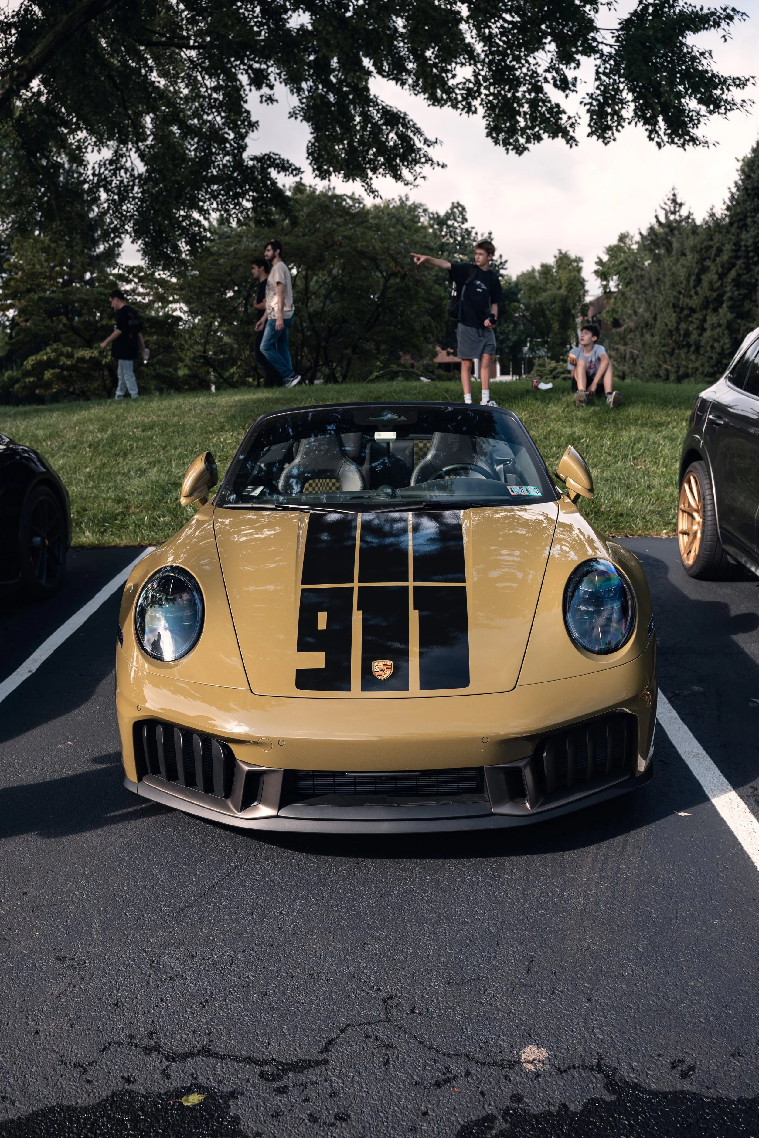 A yellow Porsche sports car with black racing stripes parked in a parking lot, with a grassy hill and trees in the background, and five people standing and sitting on the hill.