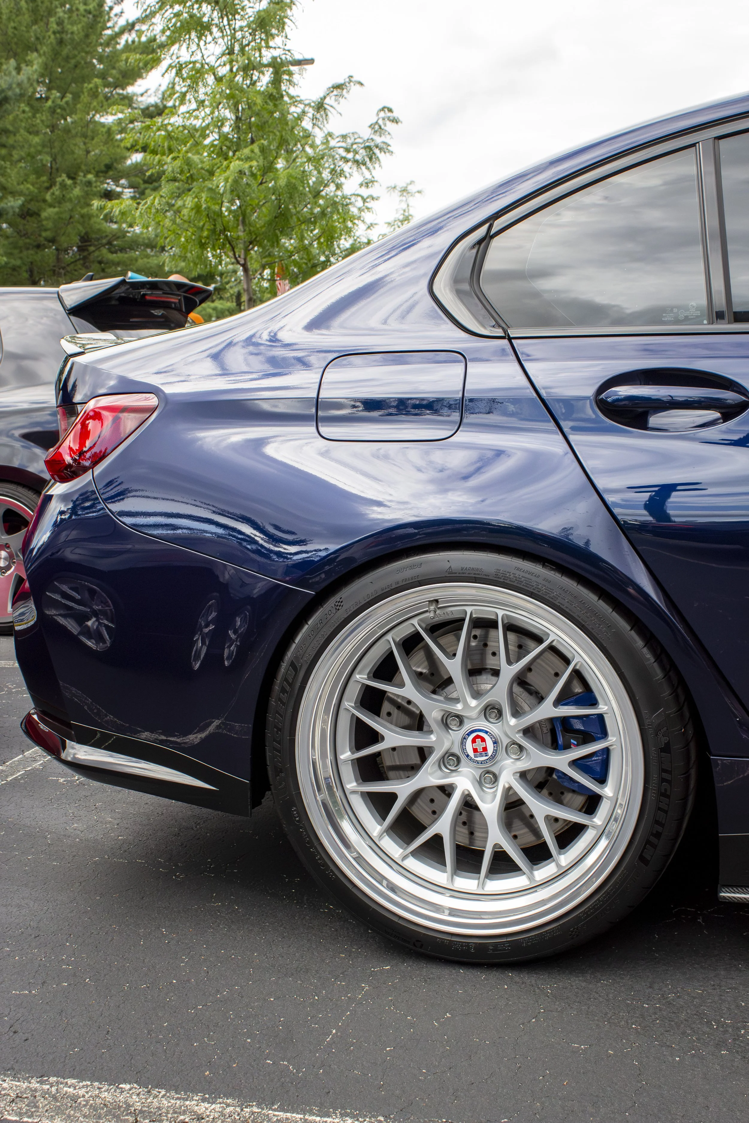 Close-up of a dark blue sports car parked on a parking lot, showing the rear quarter panel, wheel, and part of the side window, with a tree and cloudy sky in the background.