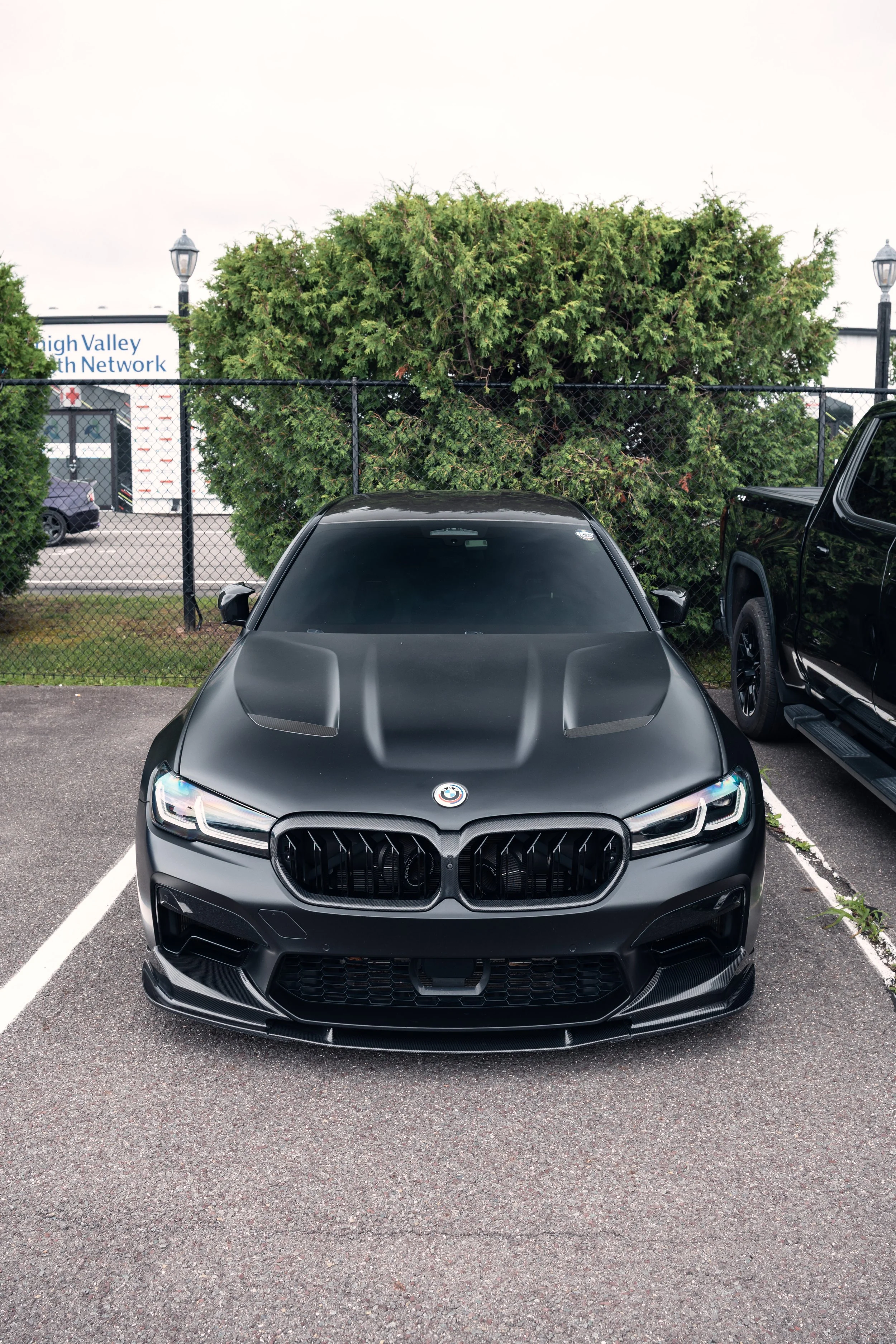 Black BMW parked in a parking lot with a chain-link fence, large bushes, and a sign that reads "High Valley Health Network" in the background.