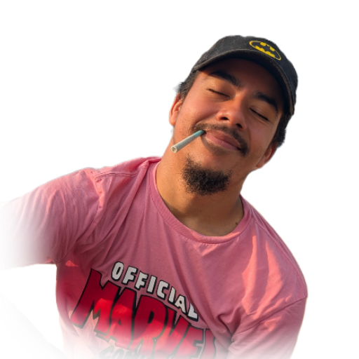 A young man with a mustache and goatee, smiling with his eyes closed, wearing a pink Marvel shirt and a black Batman cap with a yellow Batman logo, holding a cigarette in his mouth.