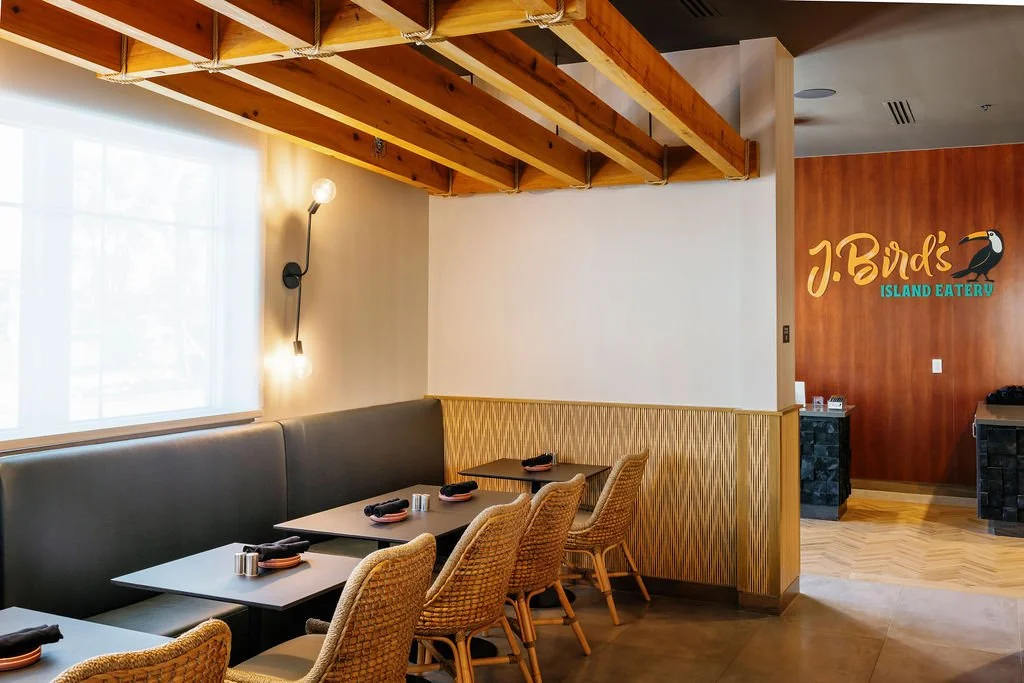 Interior of a restaurant with wooden ceiling beams, a window with white curtains, a row of tables with wicker chairs, a black wall with a J. Bird's Island Eatery sign, and a buffet area.