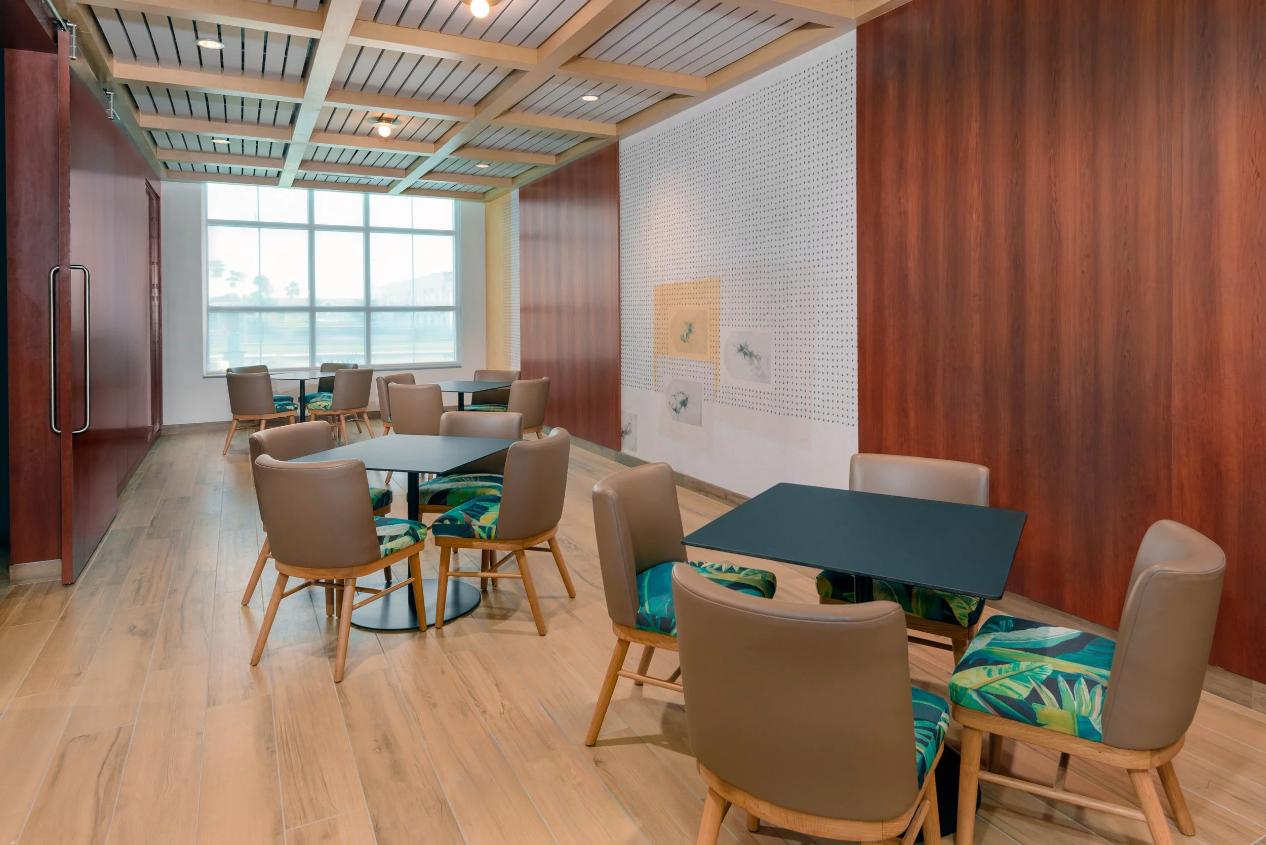 Interior of a modern cafe or lounge with wooden flooring, several tables and chairs, large window letting in natural light, decorated wall with artwork.