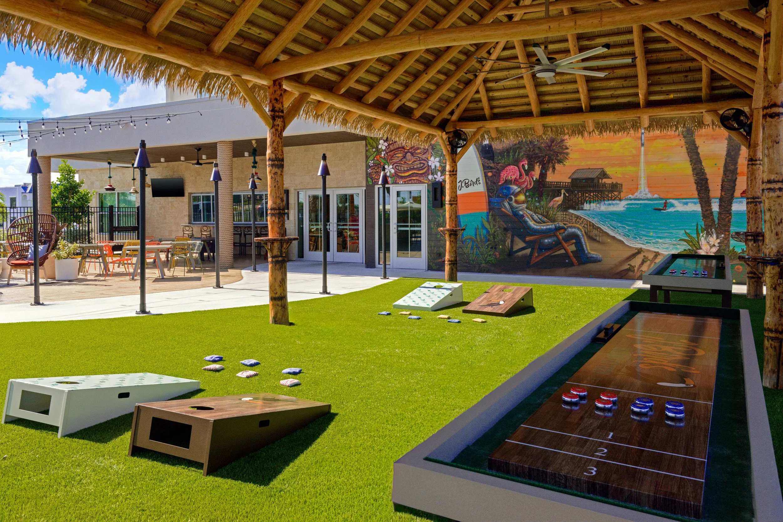 A tropical outdoor area with a thatched roof structure, painted wall mural, and lawn game setup for shuffleboard with pucks, surrounded by tables, chairs, and outdoor seating.
