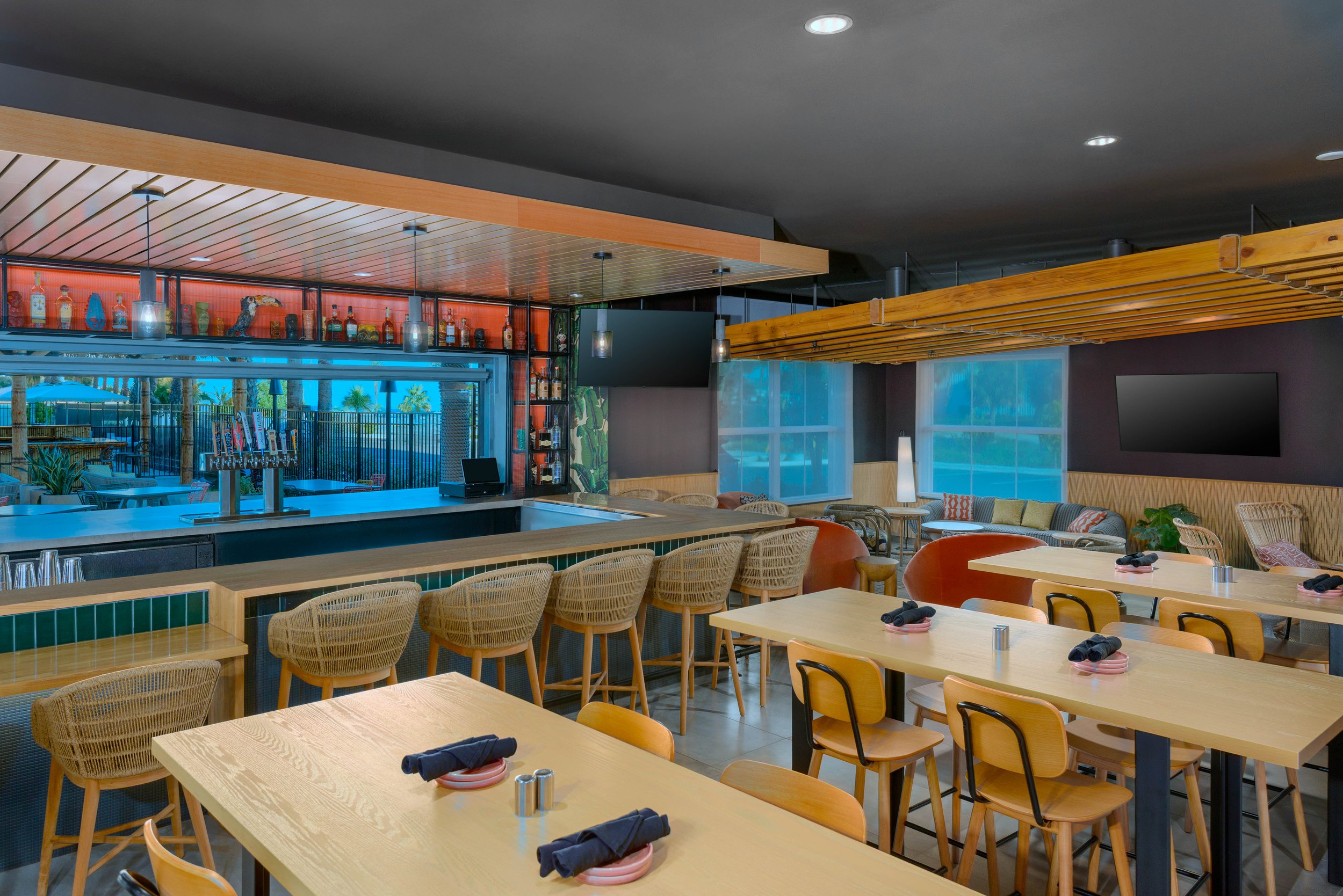 Interior of a restaurant with tables set with black napkins, rolled in a cloth, and dark plates. Wooden chairs surround the tables. A bar area features bottles on shelves, a TV, and large windows with a view of palm trees and outdoor seating.