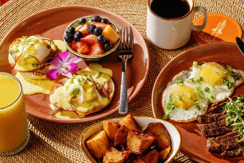 Breakfast with two eggs, roasted potatoes, fresh fruit salad, coffee, orange juice, and avocado toast, served on brown plates on a woven placemat.