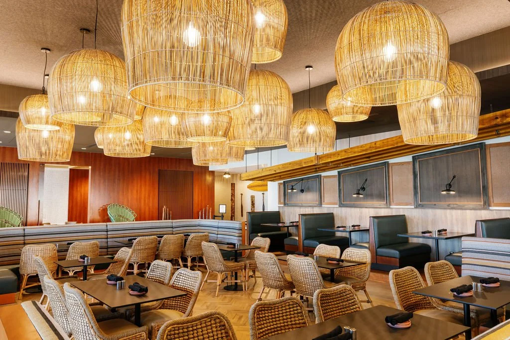 A cozy restaurant interior with bamboo pendant lights hanging from the ceiling, wicker chairs, black tables, and a mix of booth seating along the wall, decorated with framed artworks and warm wooden accents.