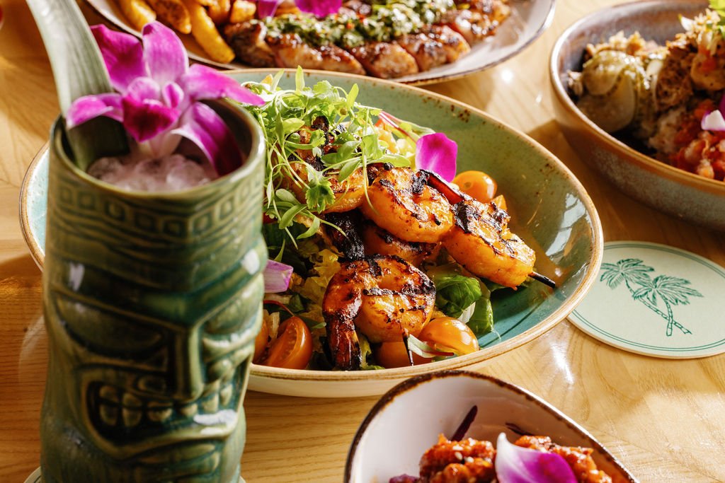 A table with various dishes, including a salad with grilled shrimp and colorful cherry tomatoes, a sushi roll, a pizza, and a drink garnished with flowers and greenery.