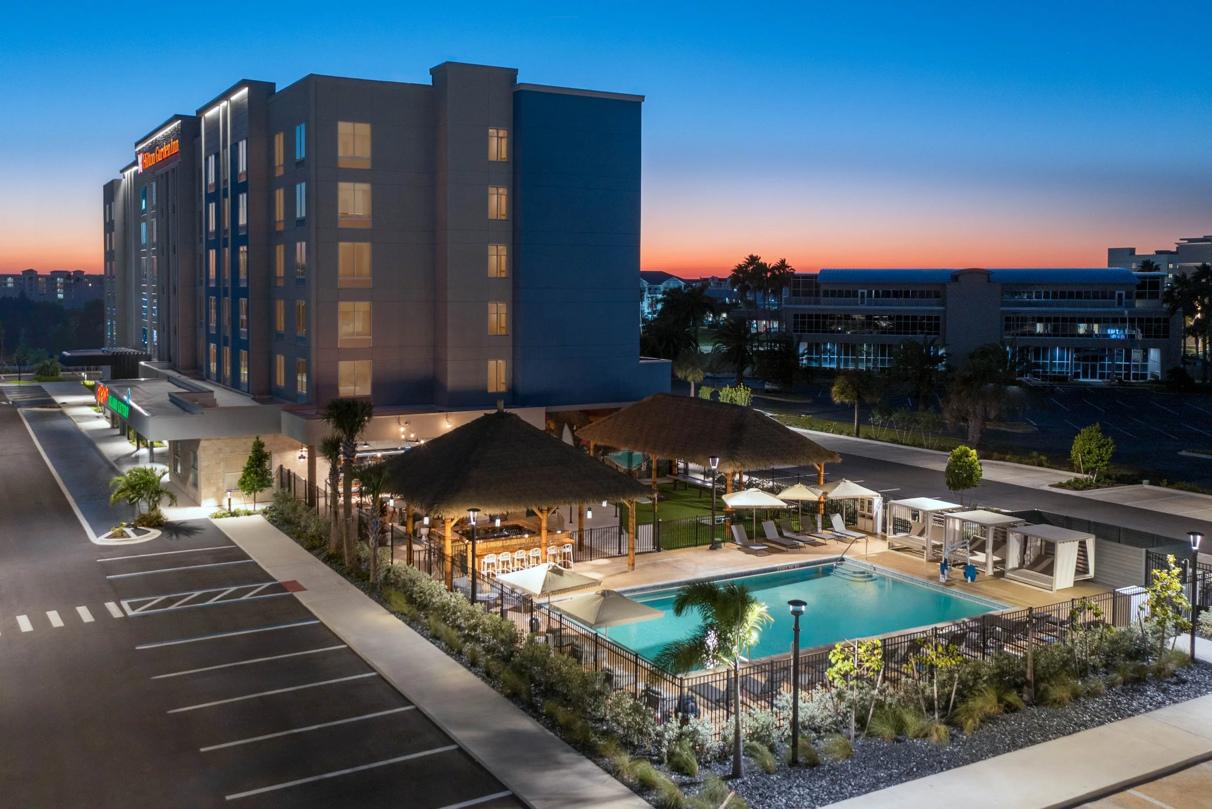 View of a Hilton Garden Inn hotel with pool and outdoor seating area at sunset. The building is multi-story with lit windows, and there are palm trees and landscaped areas around the pool and parking lot.