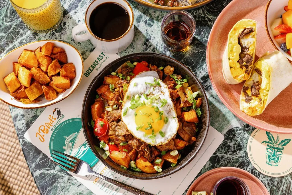 Breakfast with a bowl of beef hash topped with a fried egg, a side of roasted potatoes, two breakfast burritos filled with eggs and meat, a cup of coffee, and glasses of juice and water on a green marble table.