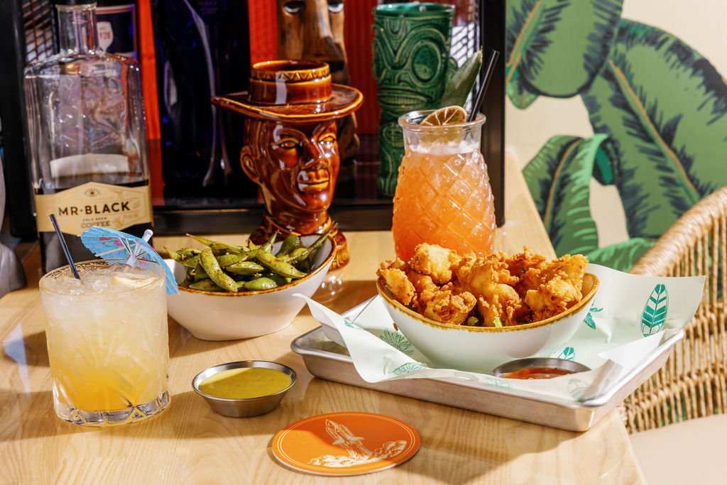 Fried chicken wings, edamame, a tropical cocktail, a lemonade drink with a paper umbrella, a yellow dipping sauce, and a large pink drink with a lemon wedge on a wooden table. There are decorative objects and leafy wall art in the background.