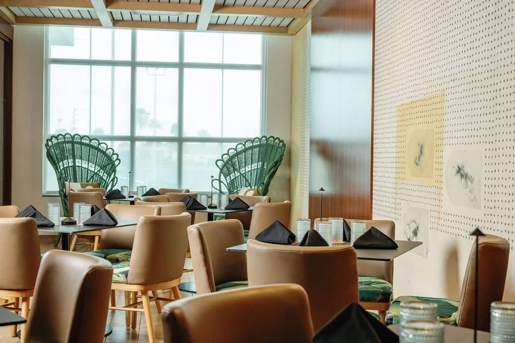 Empty restaurant with beige chairs, black napkins on tables, and large windows with outdoor views. Two green peacock chair backs visible near the window, and a dotted wall with framed drawings.