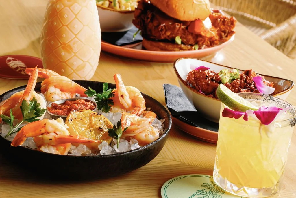 Seafood dish with shrimp, lemon, and cilantro, served on ice; two tacos with fried fish; bowl of chili; lime wedge in cocktail garnished with pink flower petal