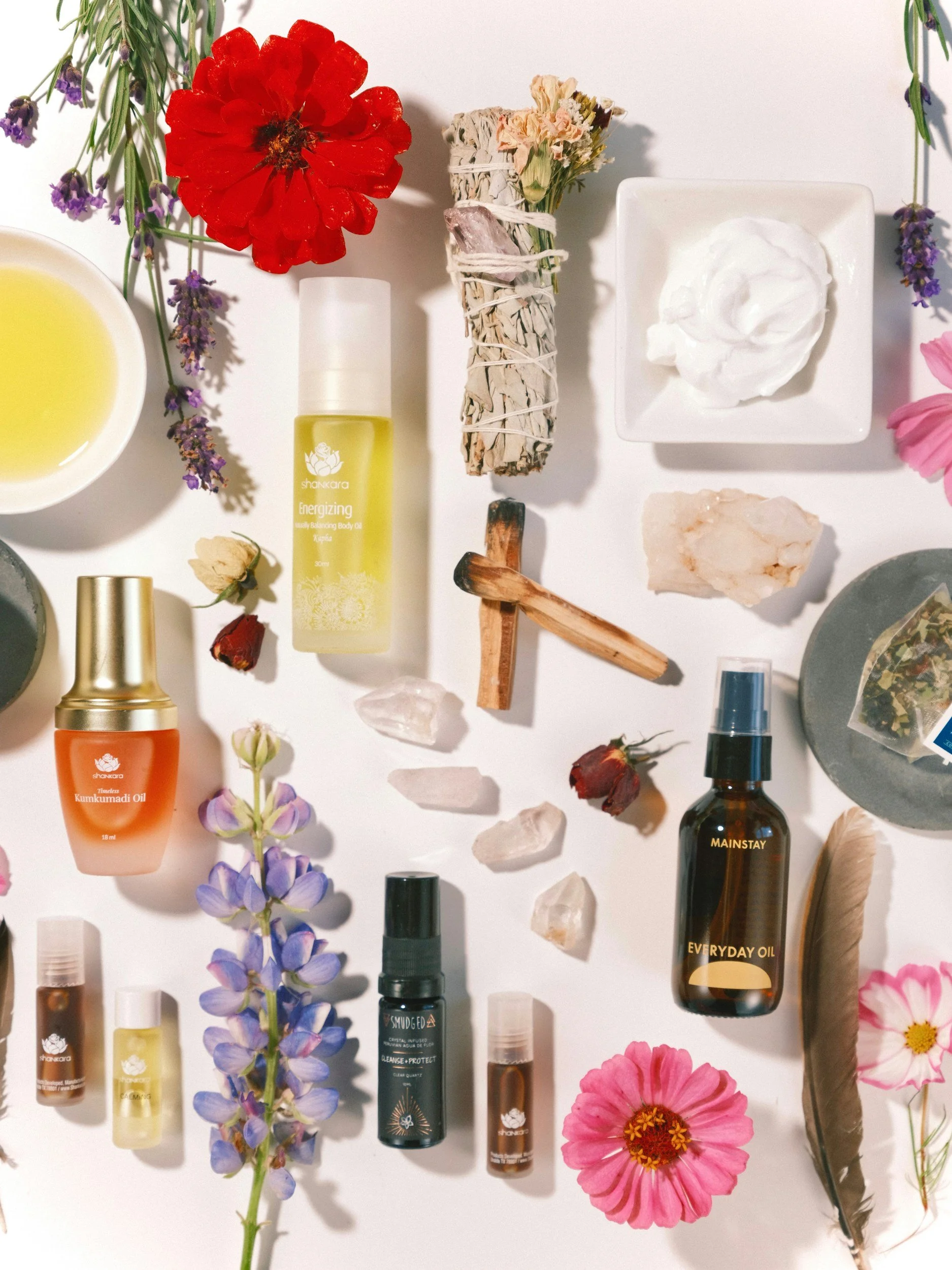 An assortment of skincare and essential oil bottles, flowers, crystals, and natural items arranged on a white background.