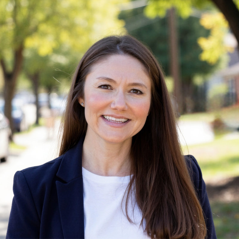 Smiling woman outdoors with long brown hair, wearing a navy blazer over a white t-shirt, on a sunny day with green trees and a sidewalk in the background.