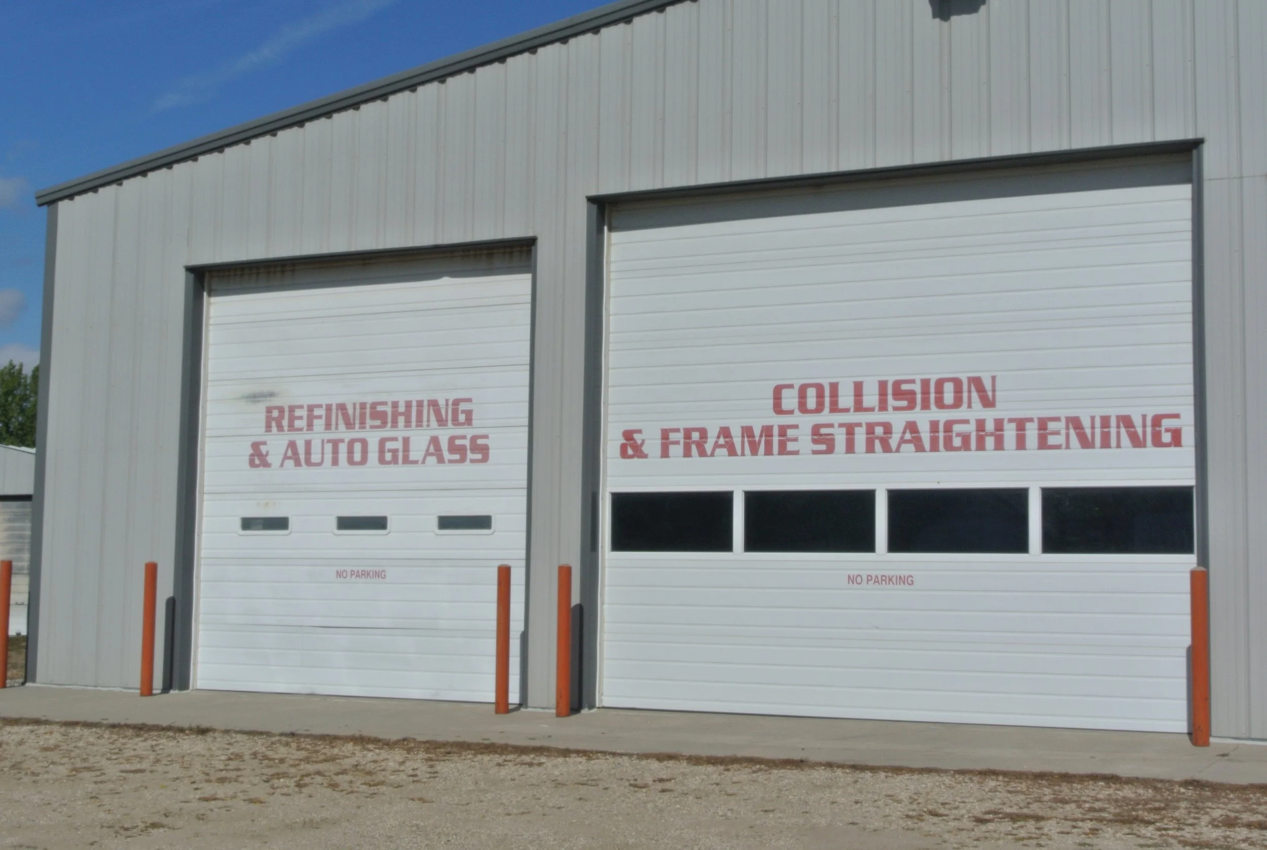 Large metal building with two closed white garage doors, each with the words "Refinishing & Auto Glass" and "Collision & Frame Straightening" written on them in red. Orange safety posts are in front, and the ground is unpaved with dirt and gravel. The sky is blue with some clouds.