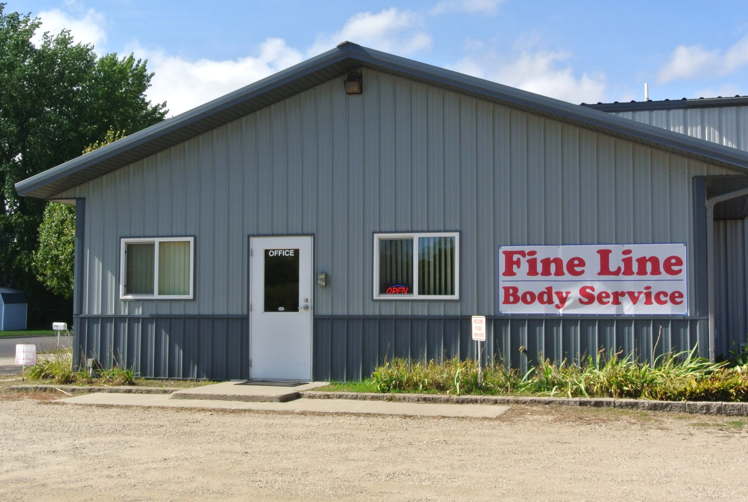 Gray metal building with a sign that reads 'Fine Line Body Service', a glass door labeled 'Office', and a small 'Open' sign in the window. The office building has two windows, and there's a small lawn in front with plants.