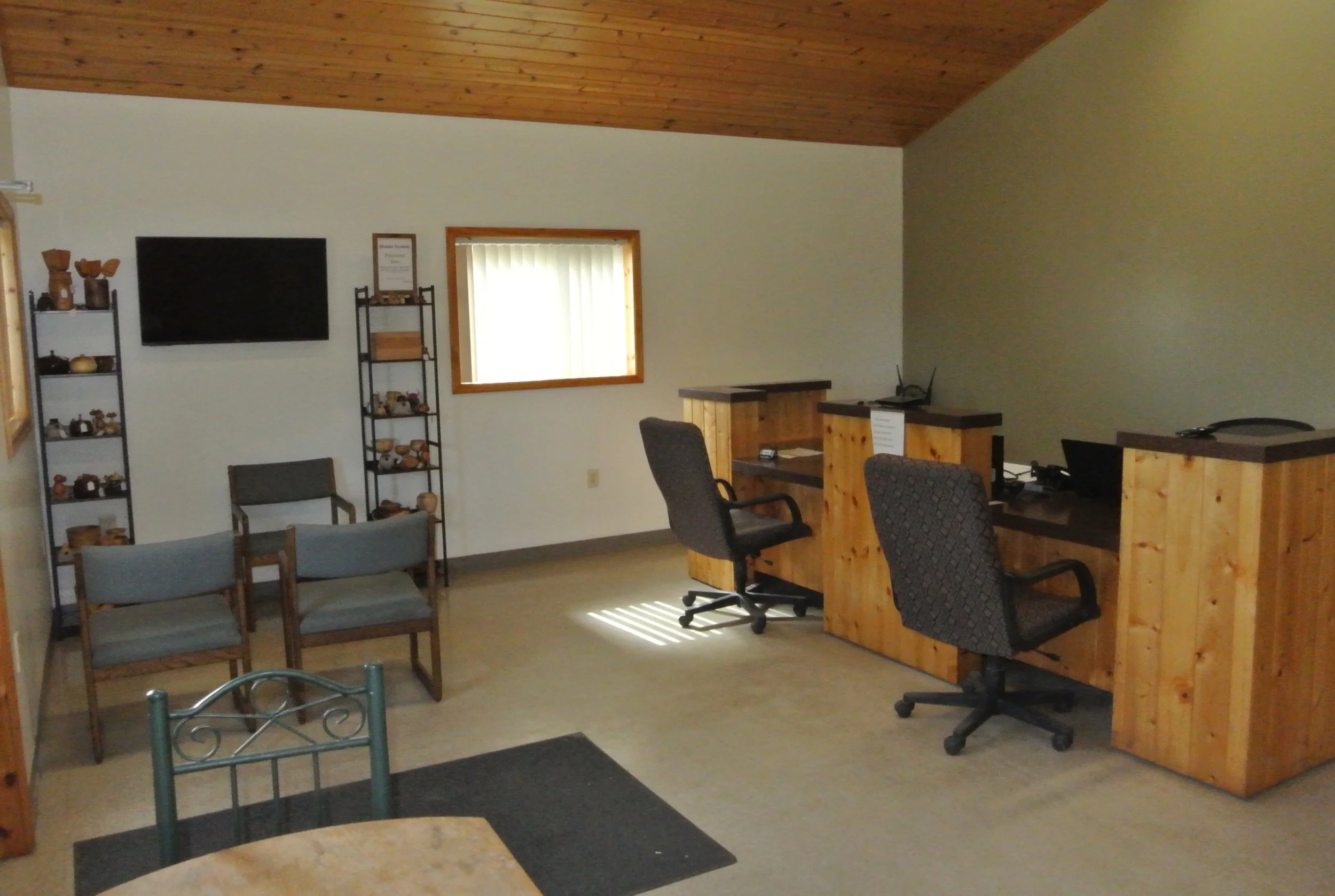 Interior of a room with a wooden vaulted ceiling, cream-colored walls, and a window with vertical blinds. The room features shelves with pottery, a flat-screen TV, a small counter with chairs, and office chairs behind a wooden partition.