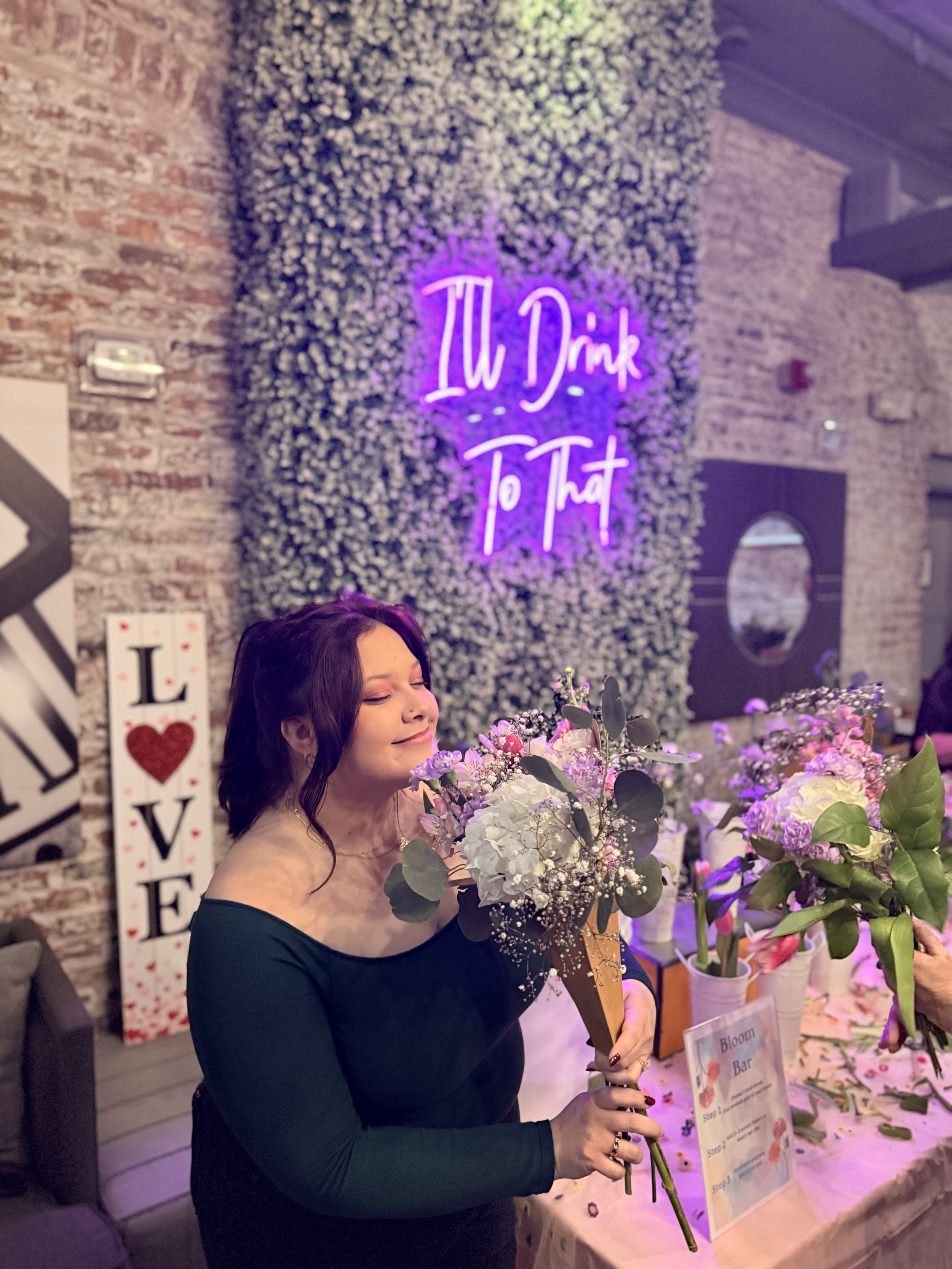 A woman with dark hair is holding a large bouquet of mixed flowers, including white and pink blossoms, at a floral display or event. There is a sign on the table that says 'Bloom Bar' and a neon sign in the background that says 'I'll Drink To That.' The setting appears to be inside a decorated venue with brick walls and floral arrangements.