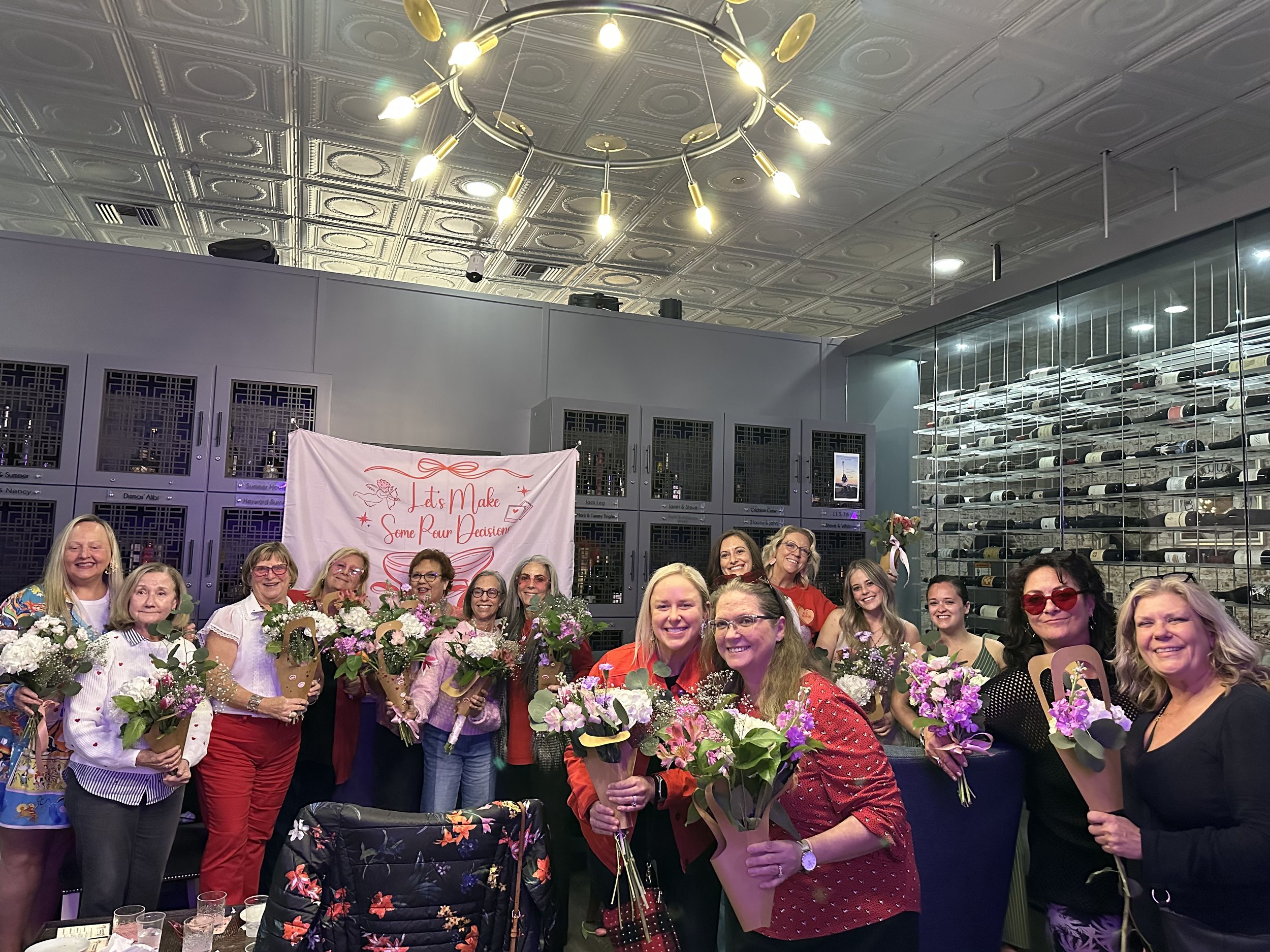 A group of smiling women holding flowers at a celebration event in a room with a decorative ceiling, a chandelier, and a wall of wine bottles.