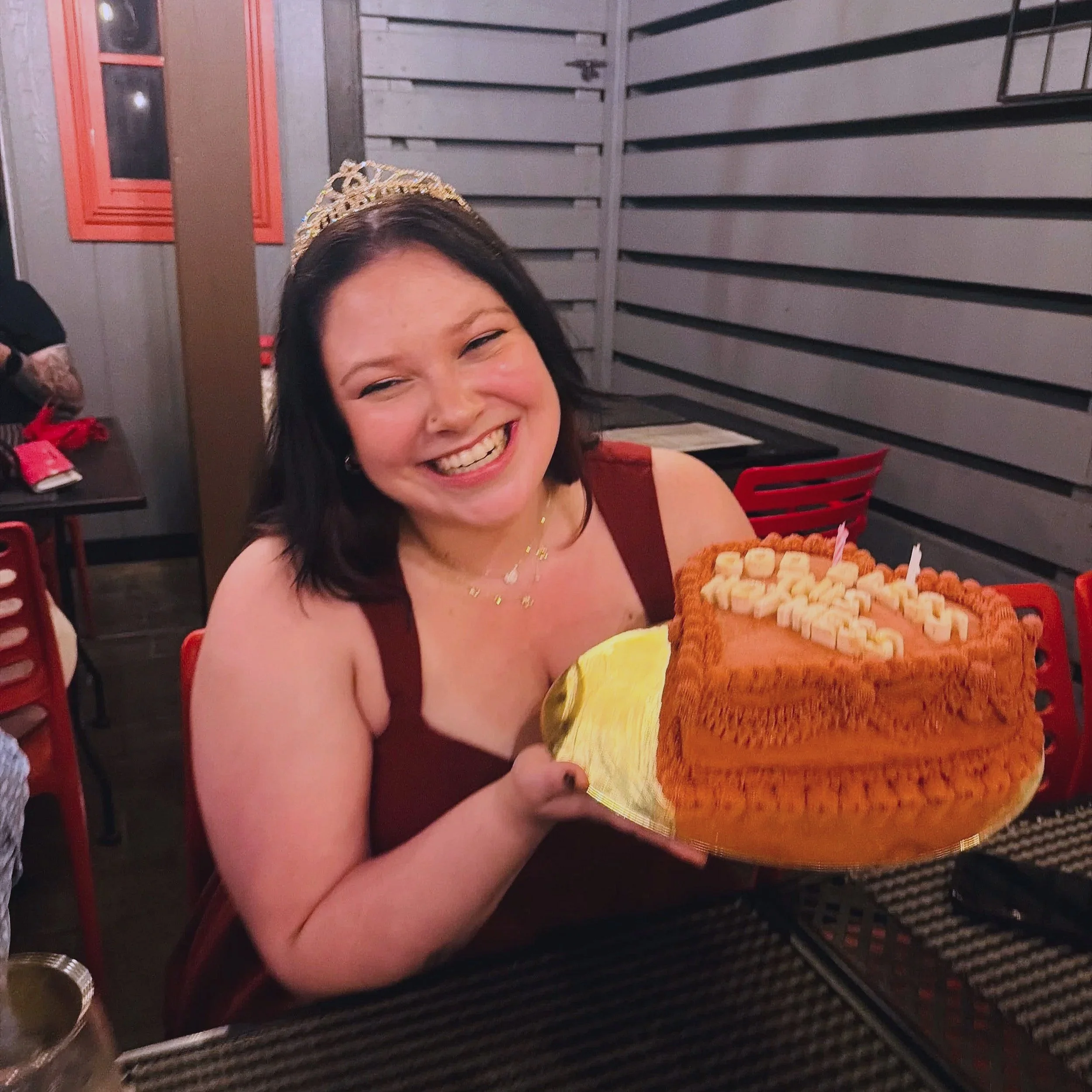 A woman smiling while holding a birthday cake with lit candles in a restaurant. She is wearing a tiara and a maroon top, and the background shows metal wall panels and red chairs.