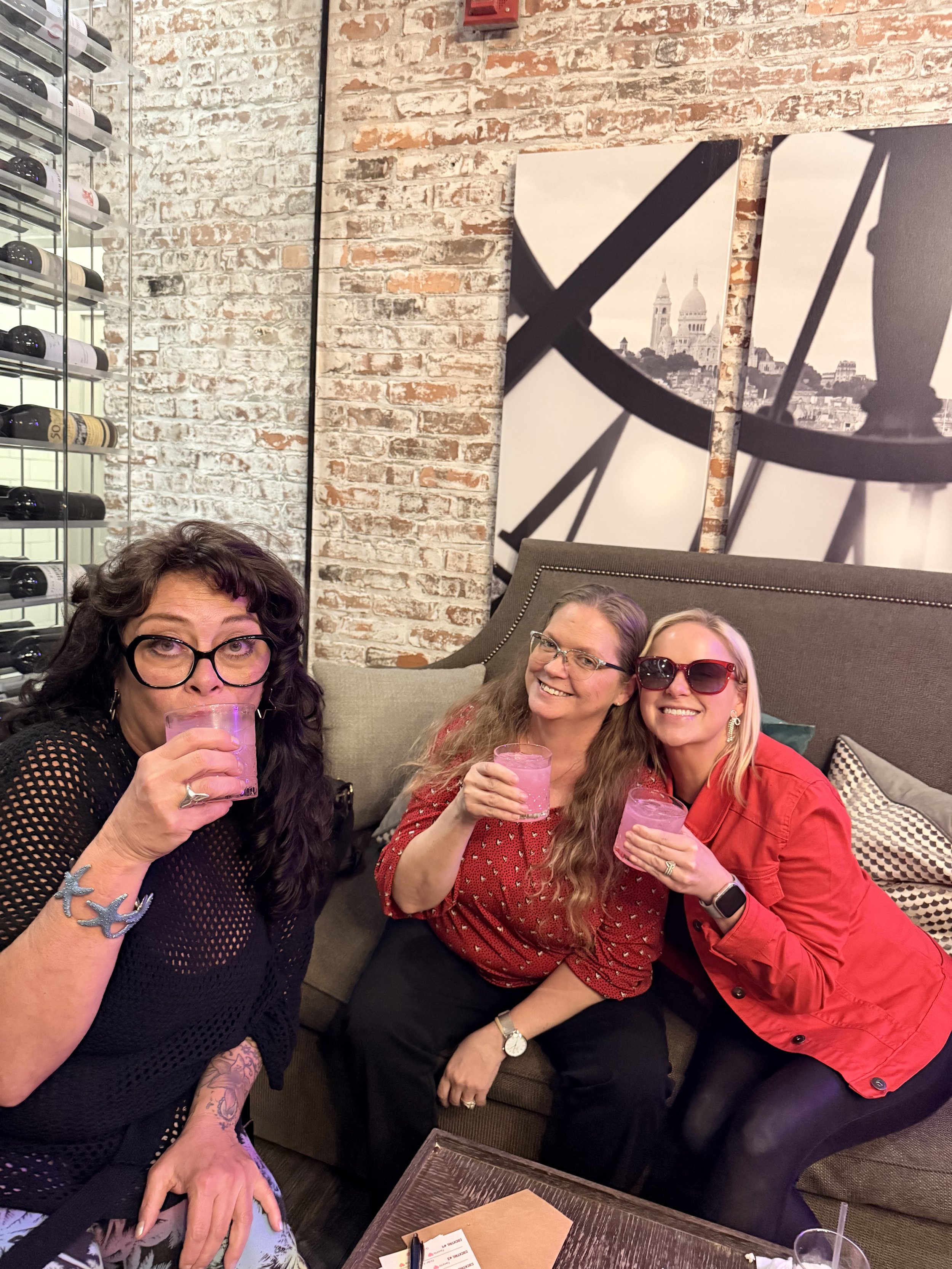 Three women sitting close together on a sofa, holding pink drinks and smiling at the camera in a cozy indoor setting with exposed brick walls and large black-and-white cityscape artwork.