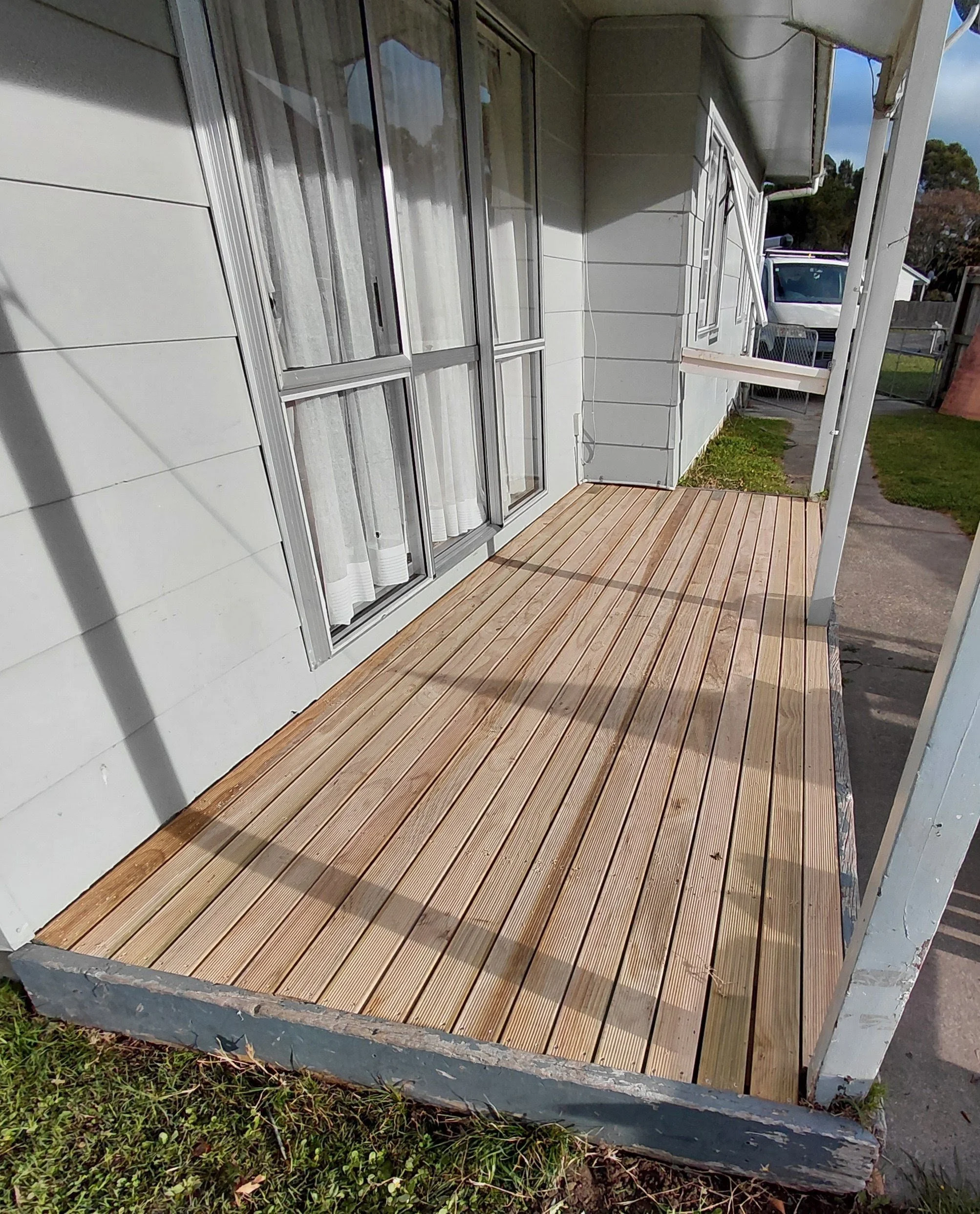 A small porch with a wooden floor in front of a house with large, sliding glass doors. A white curtain hangs inside the house, and sunlight casts shadows on the house's exterior walls. A white vehicle is visible in the driveway, and the surrounding y
