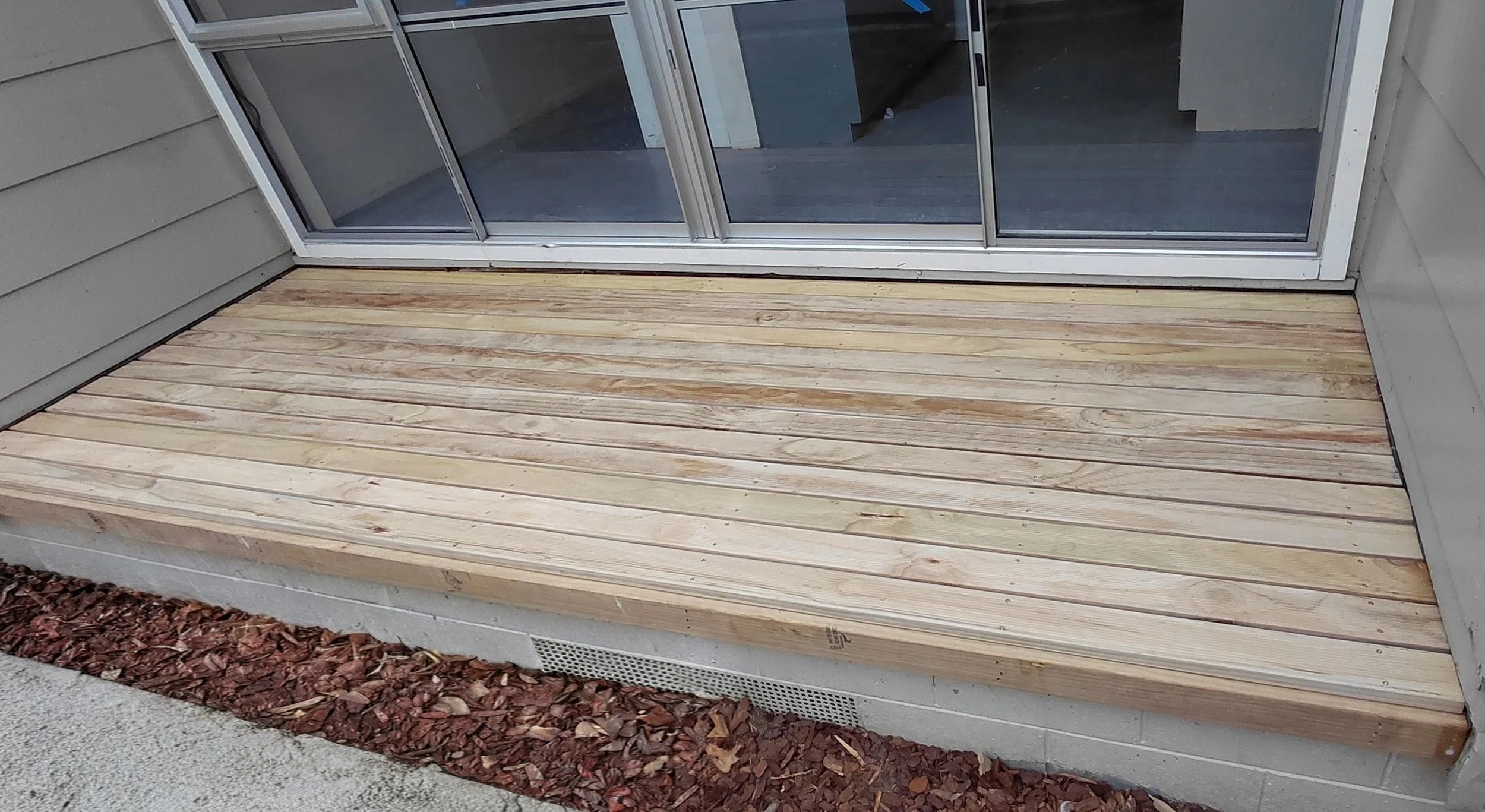 Newly installed wooden deck outside sliding glass door with gray siding and a vent or speaker below the deck, surrounded by mulch and concrete.