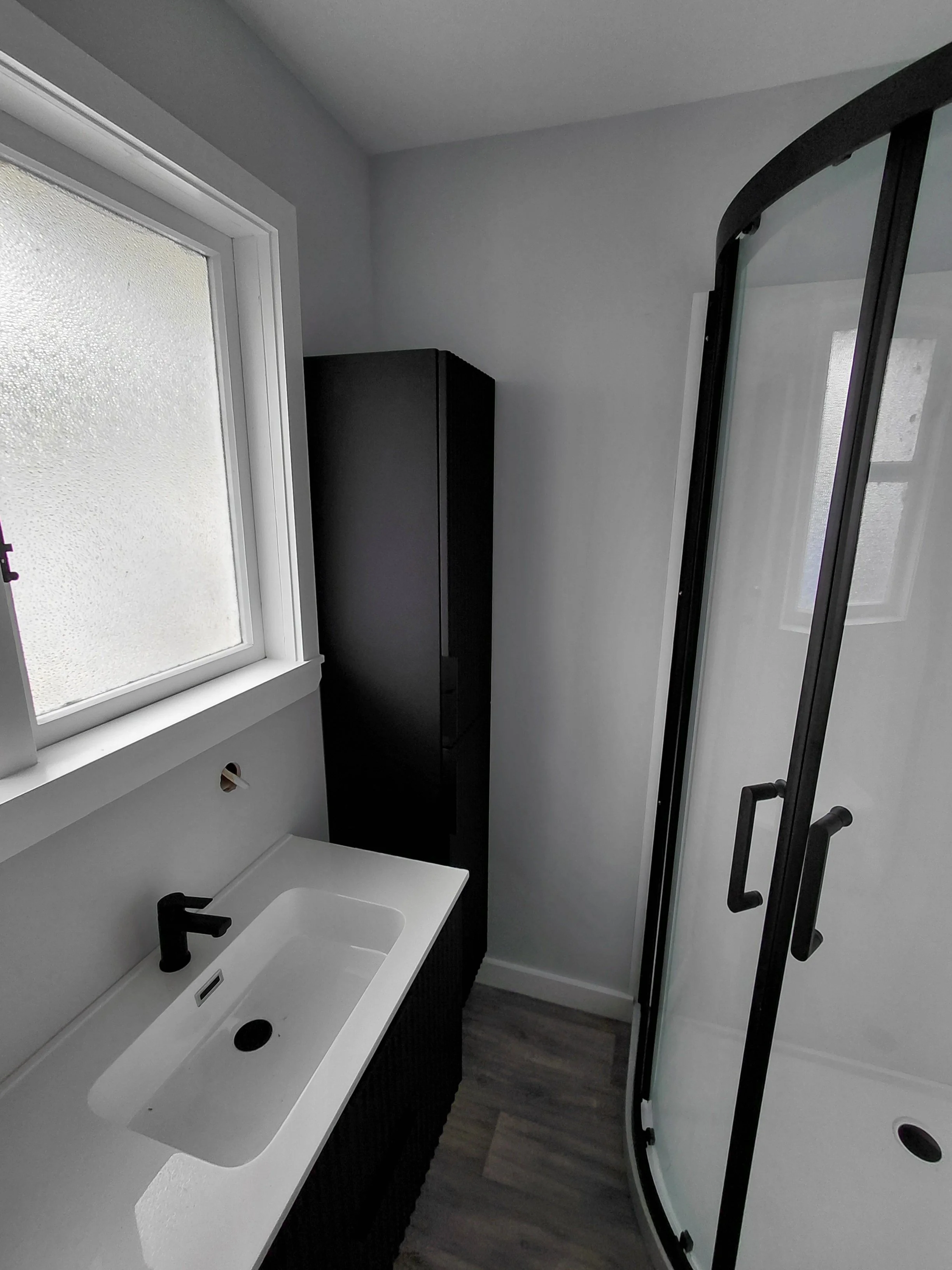 Small bathroom with frosted window, white sink with black faucet, black cabinet, and curved glass shower door with black handles.