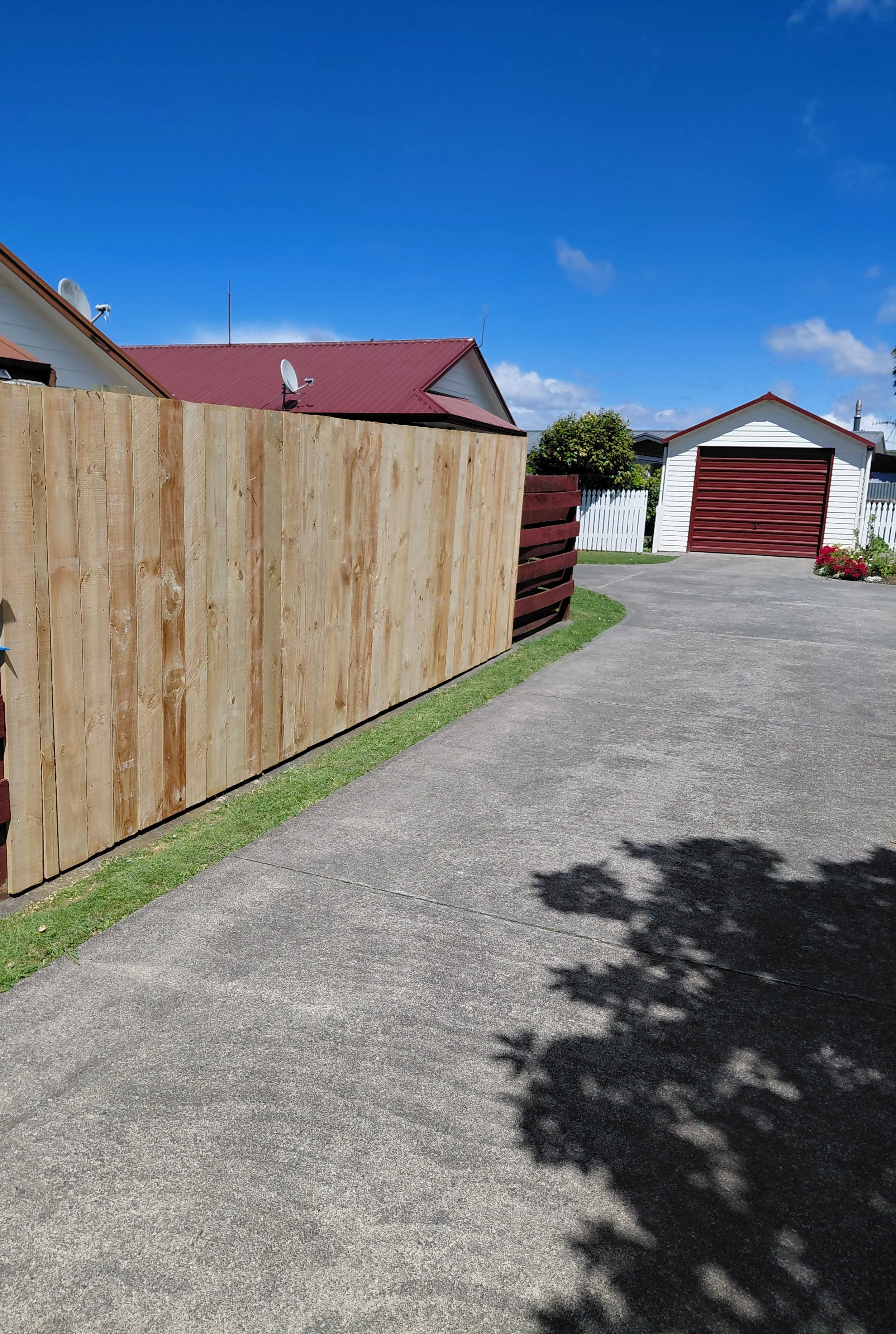 A suburban backyard with a concrete driveway, a wooden fence, a white shed with a red garage door, and a house with a red roof in the background. Clear blue sky with a few clouds.