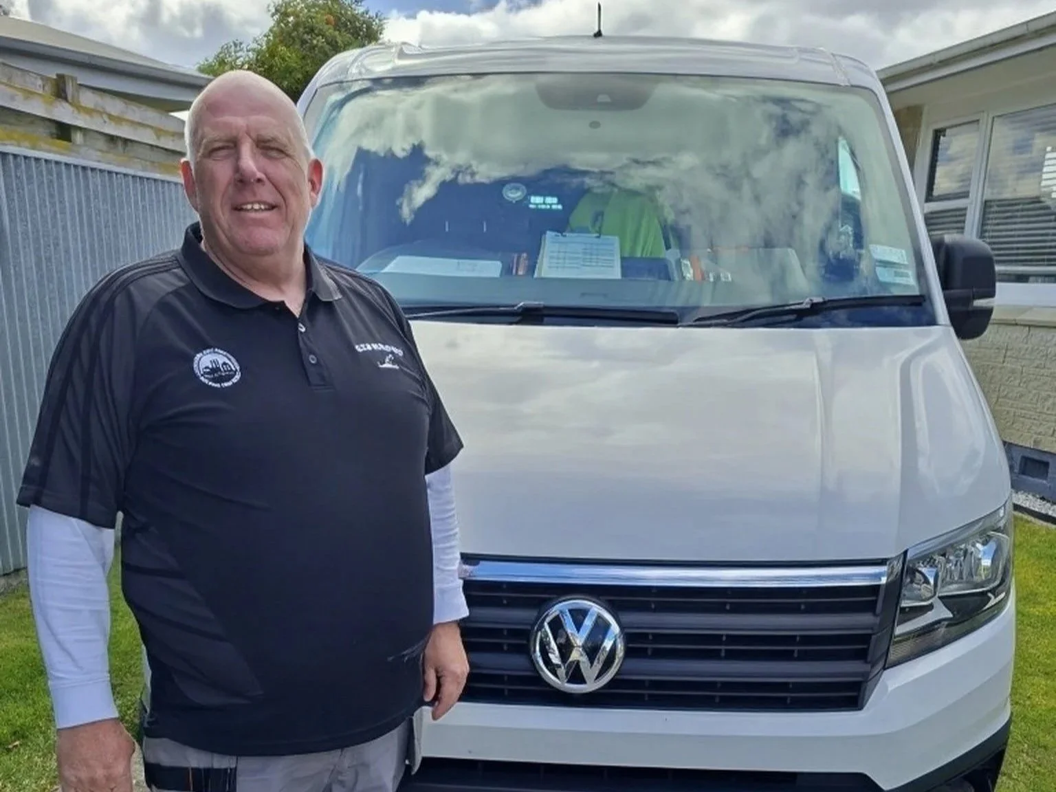 A smiling man standing in front of a white Volkswagen van in a backyard, with a house and trees in the background.