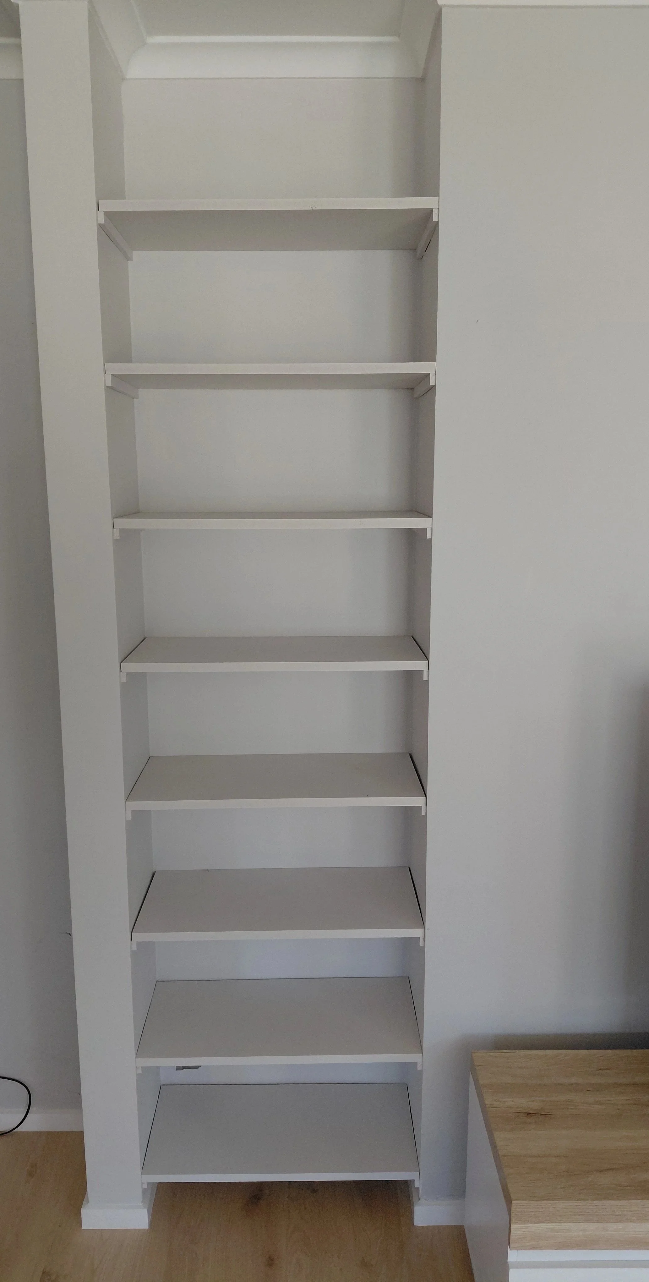 Empty white bookshelf with five shelves, next to a small wooden table or cabinet.