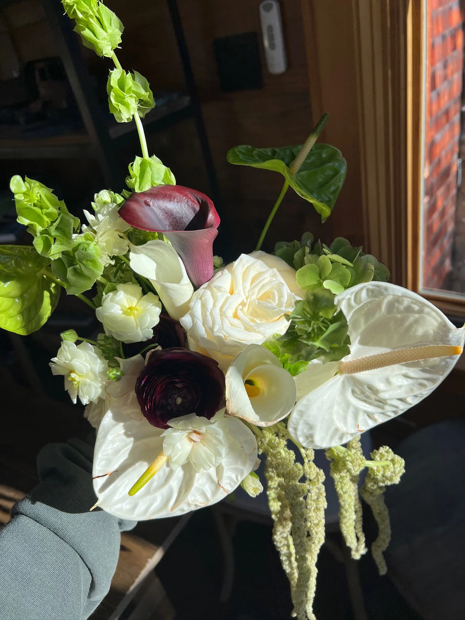 A floral arrangement with white roses, green hydrangeas, dark purple ranunculus, white anthuriums, a pink calla lily, and an anthurium leaf, set against a wooden wall background near a window.