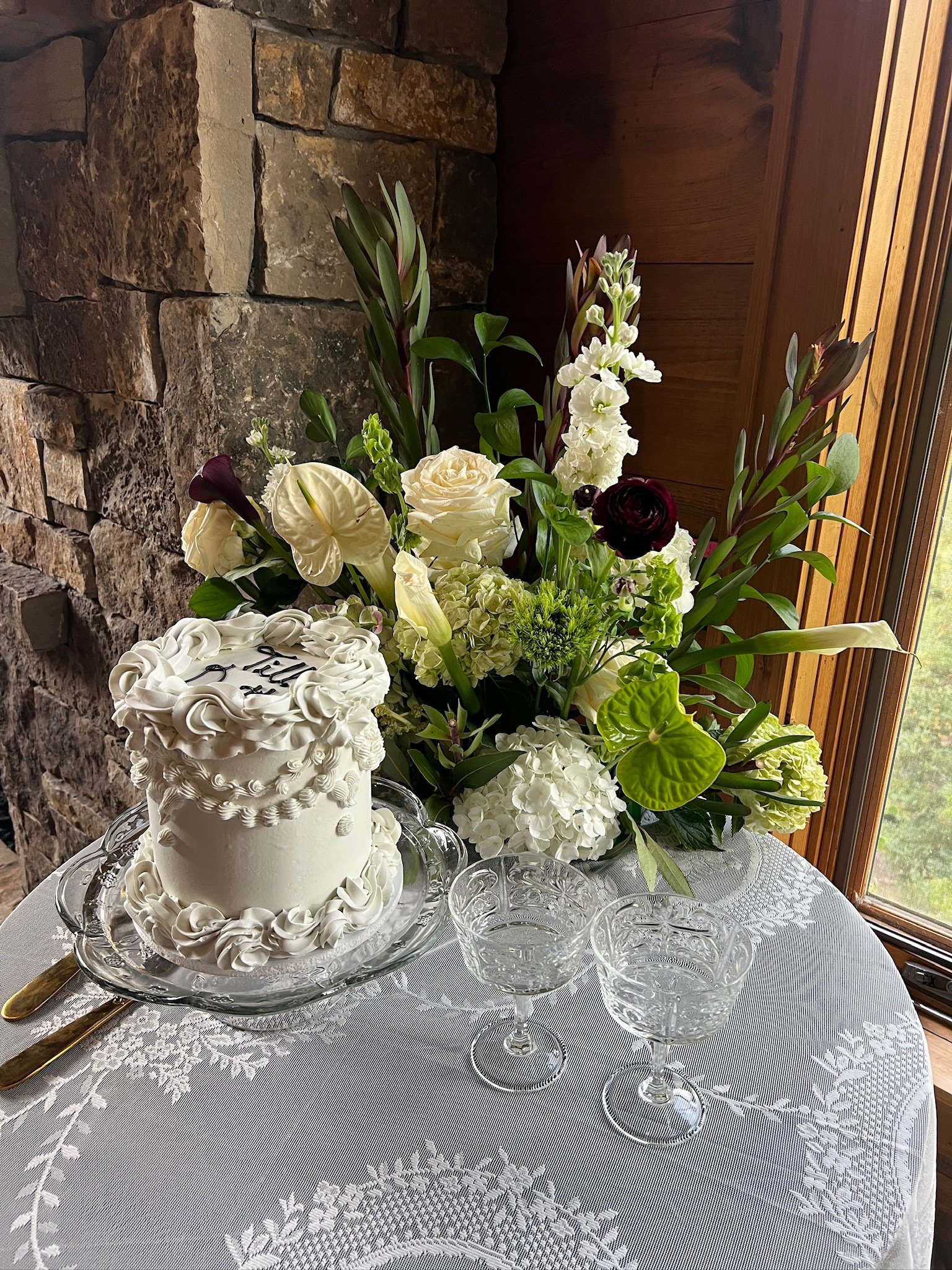 A wedding cake with white icing and decorative roses, placed on a glass tray on a lace tablecloth. Behind the cake is a large bouquet of white, purple, and green flowers, set against a stone wall with a wooden window to the right.