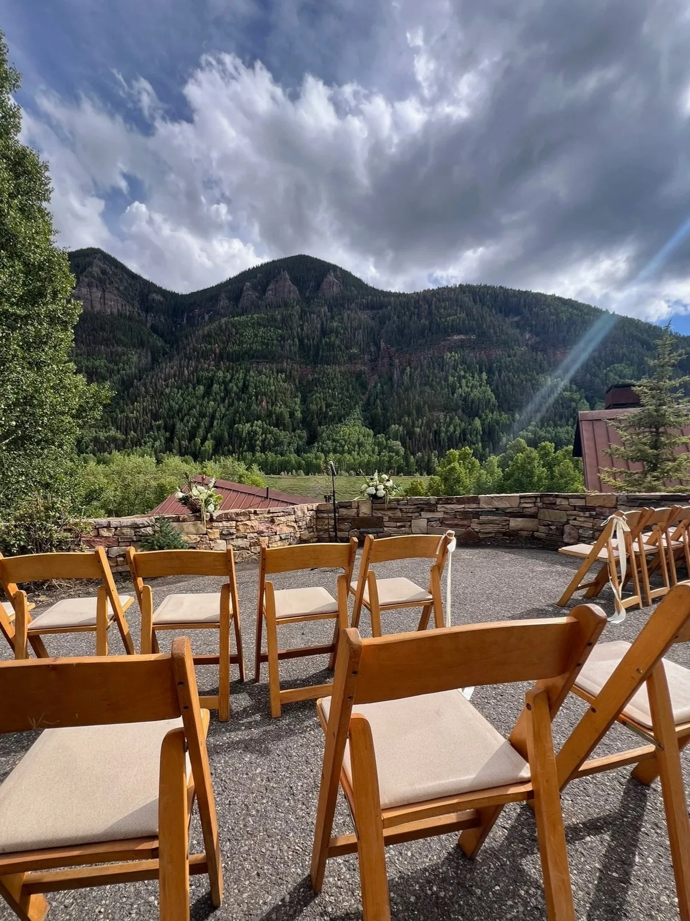 A scenic outdoor patio with wooden chairs arranged on a gravel surface, overlooking a lush green mountain range under a partly cloudy sky.