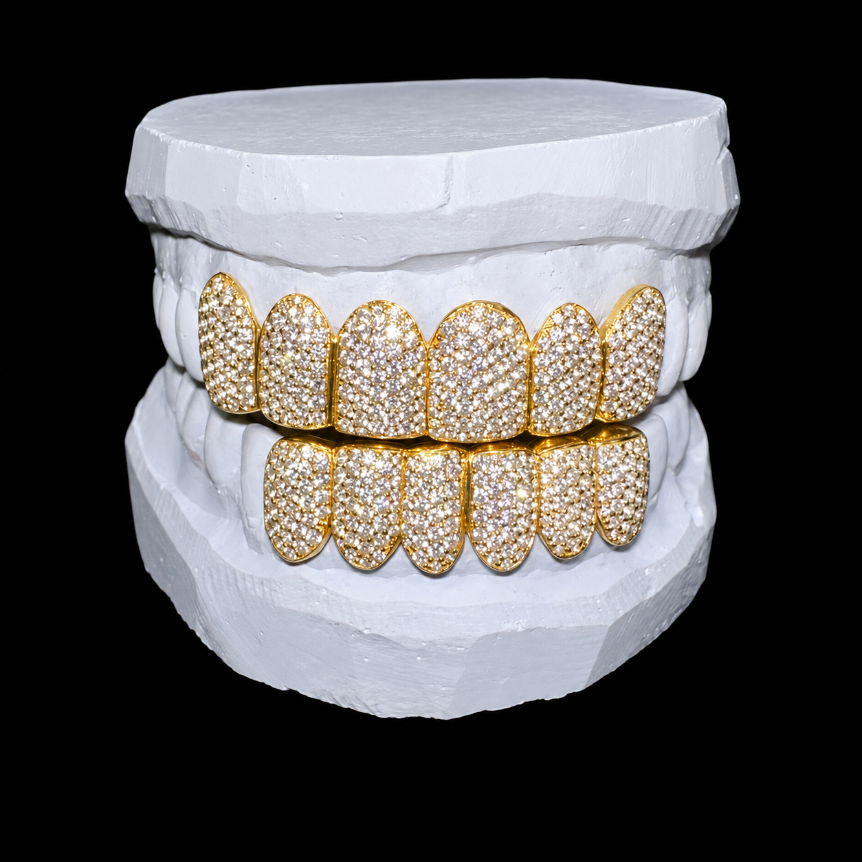 A set of gold grillz encrusted with diamonds on a white dental mold, reflected on a black surface.