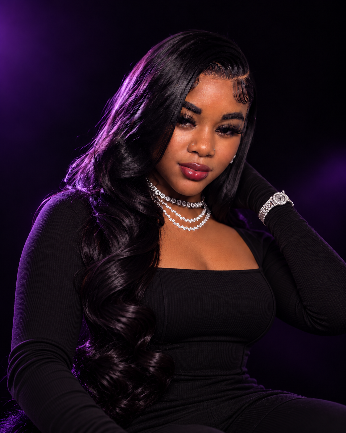 Portrait of a young woman with long, wavy black hair, wearing a black long-sleeve top, layered necklaces, a watch, and makeup, against a dark background with purple lighting.