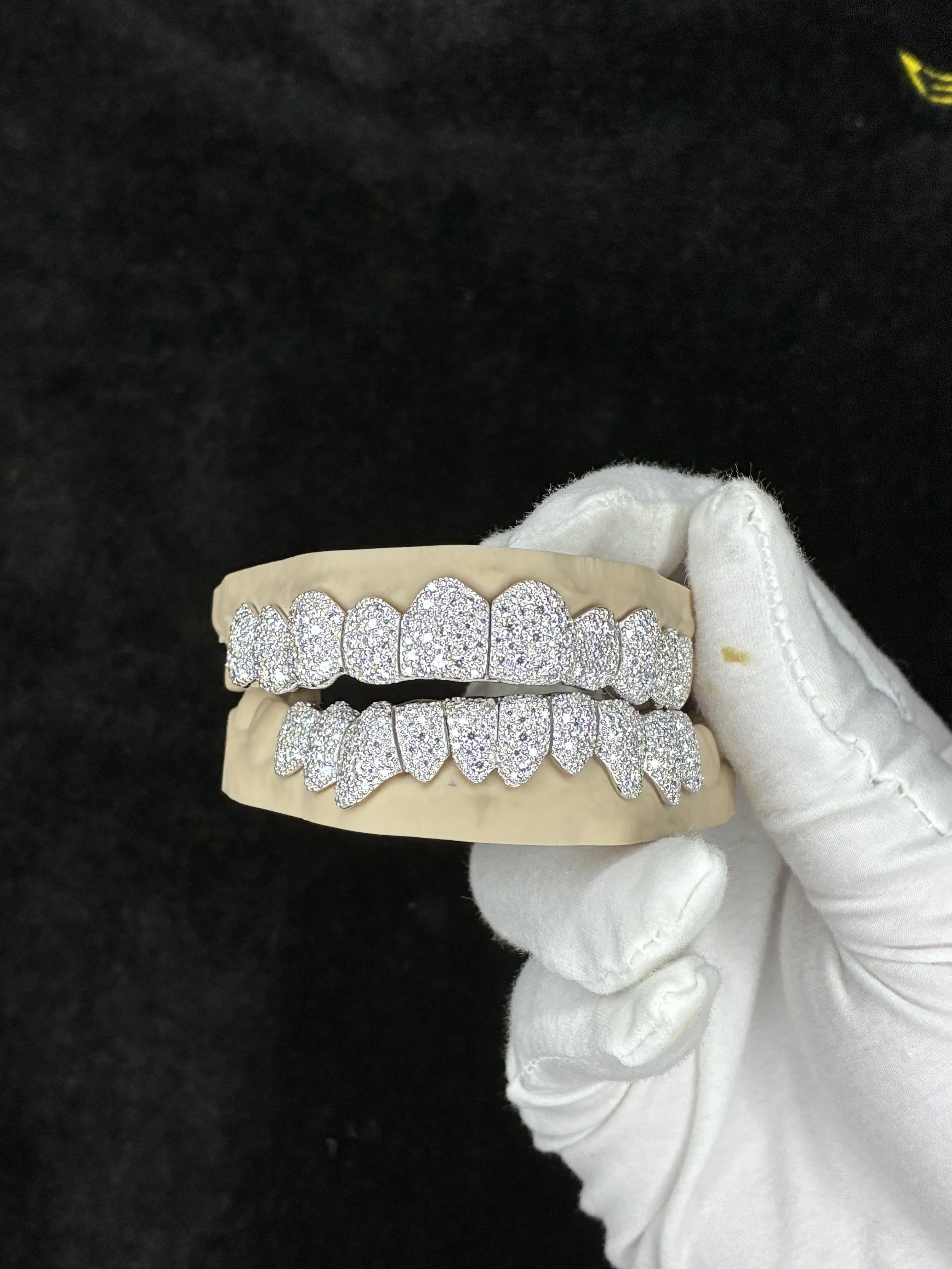 A gloved hand holding a set of shiny, diamond-encrusted grillz on a beige display tray against a black background.