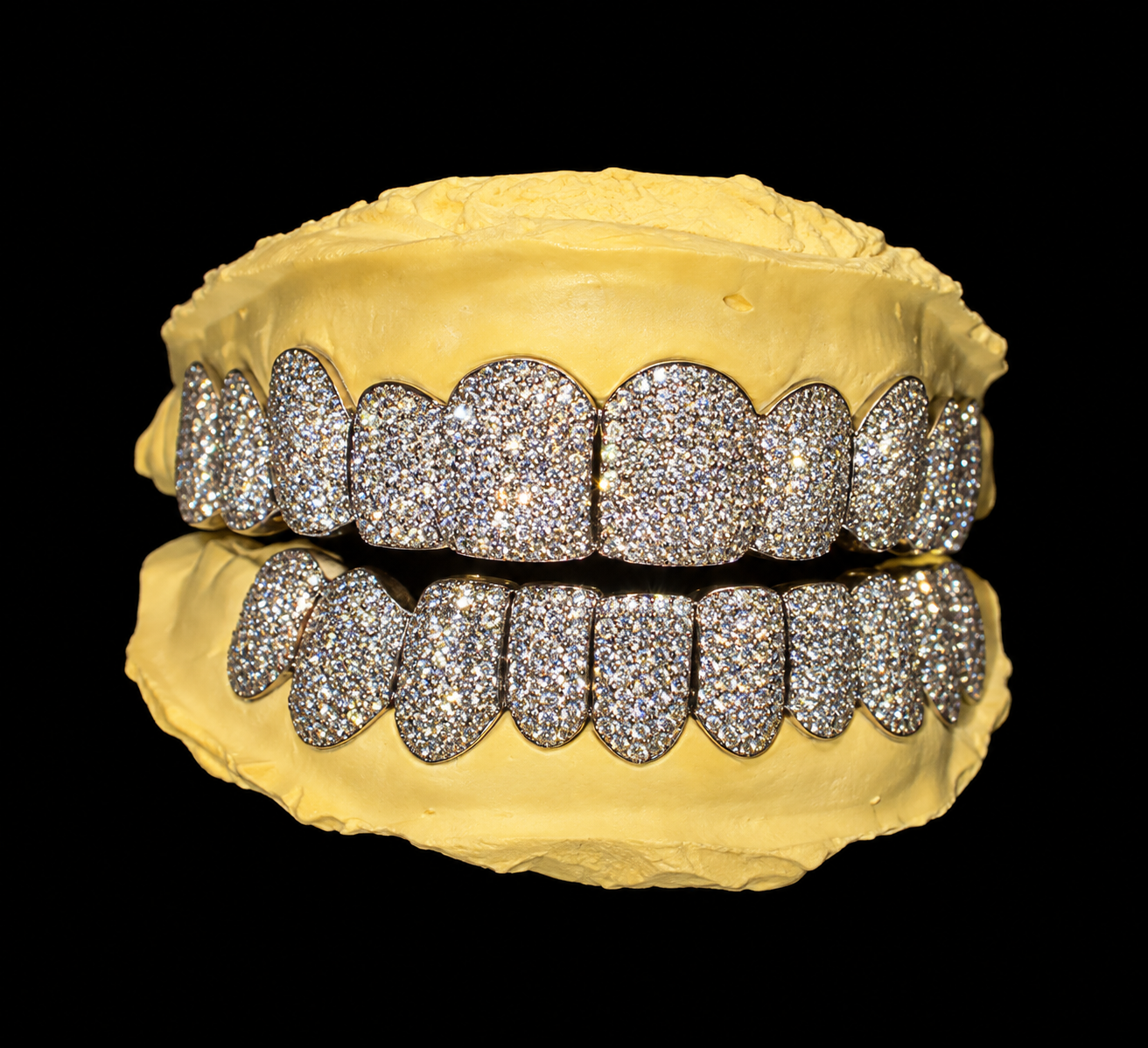 A set of diamond-studded jewelry in the shape of teeth on a yellow material, reflected on a black surface against a black background.