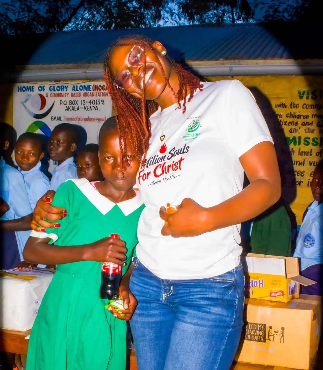 A woman wearing glasses and a t-shirt with the words 'Milton Souls for Christ' is smiling and arm around a young girl holding a bottle of Coca-Cola and a small item. Several children are in the background, some watching and some looking at the camera