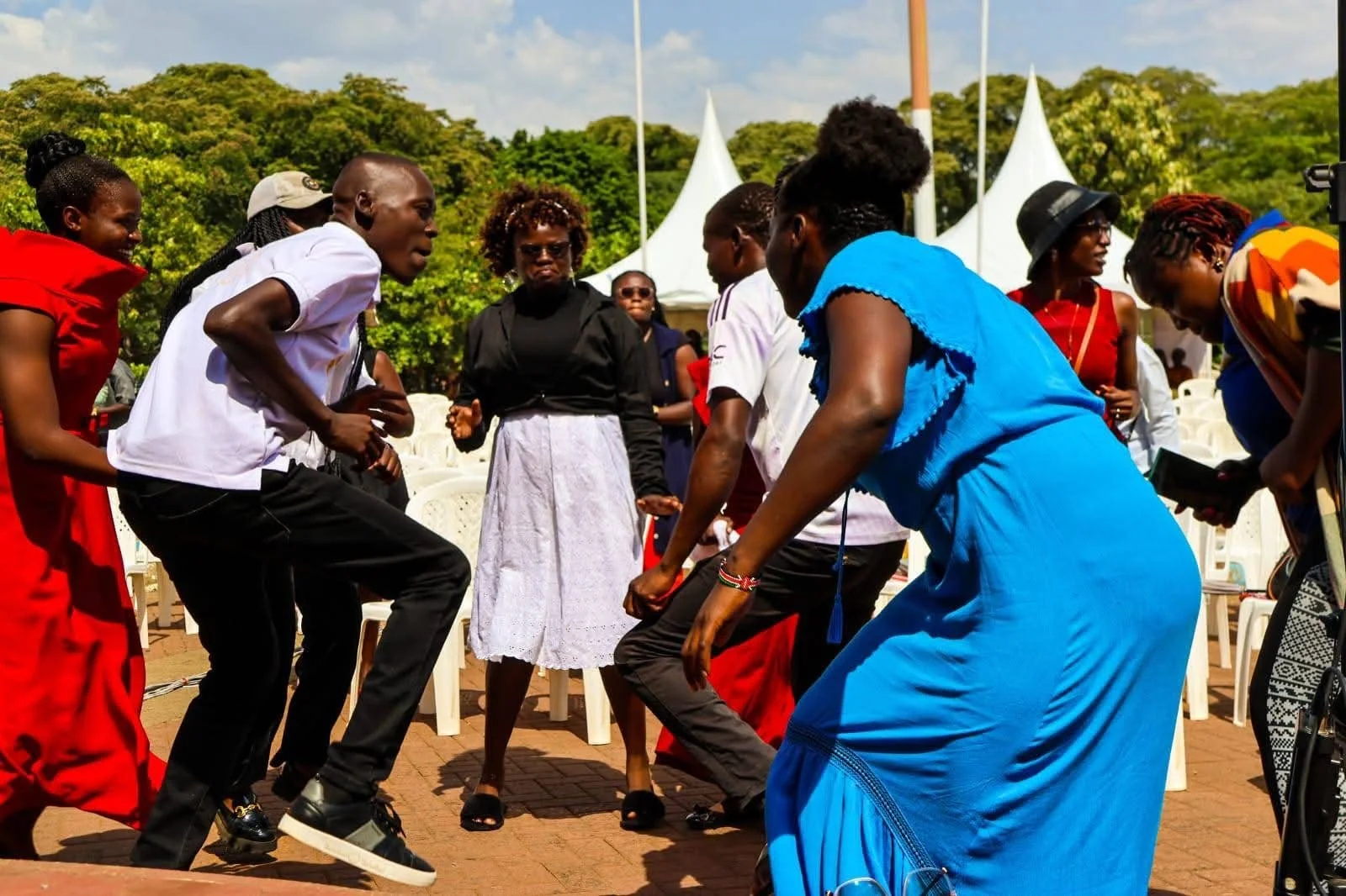 Group of people dancing outdoors on a sunny day, with trees and tents in the background.