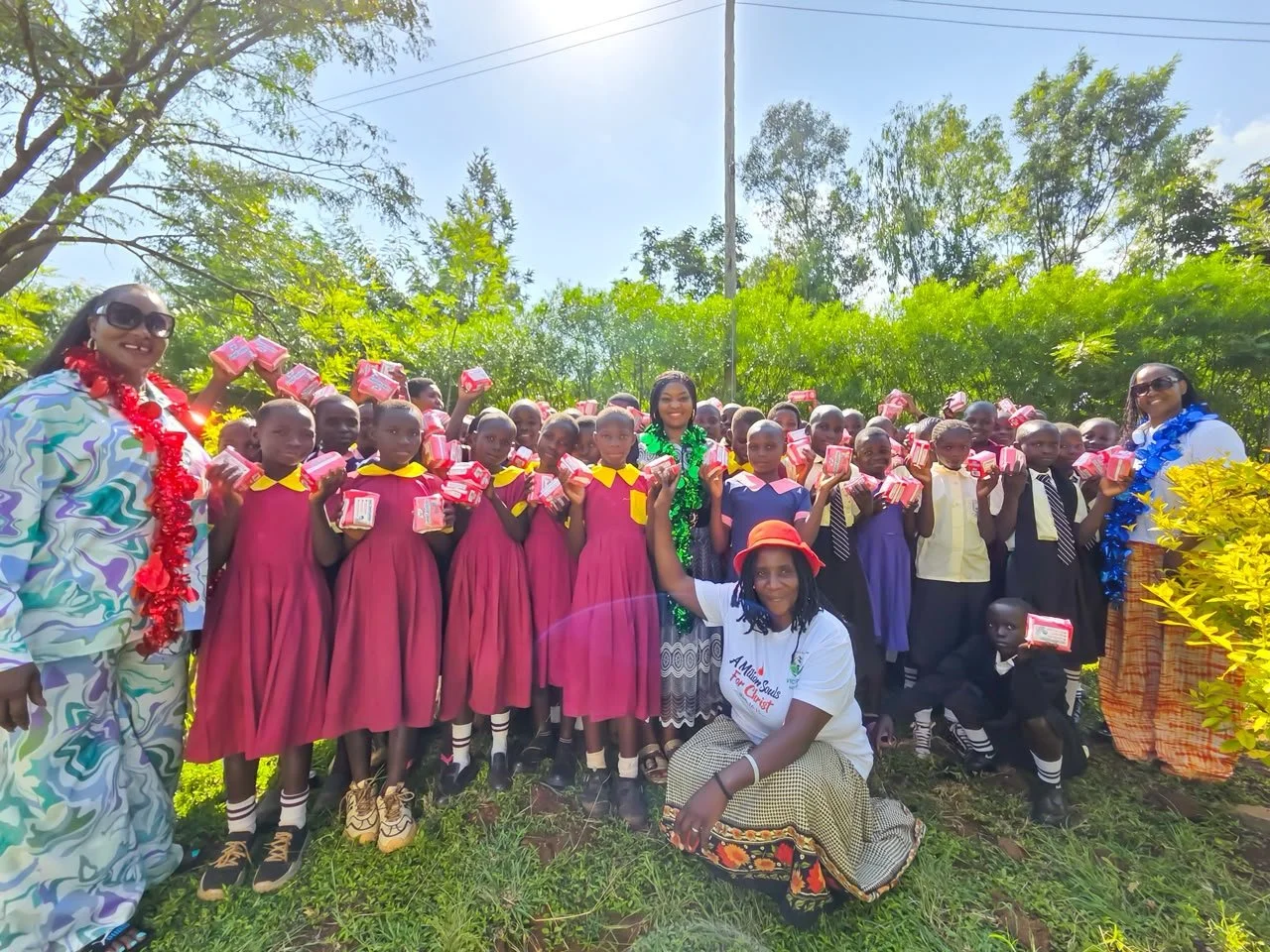 Group of children and adults outdoors, holding gifts, smiling, with greenery in the background.