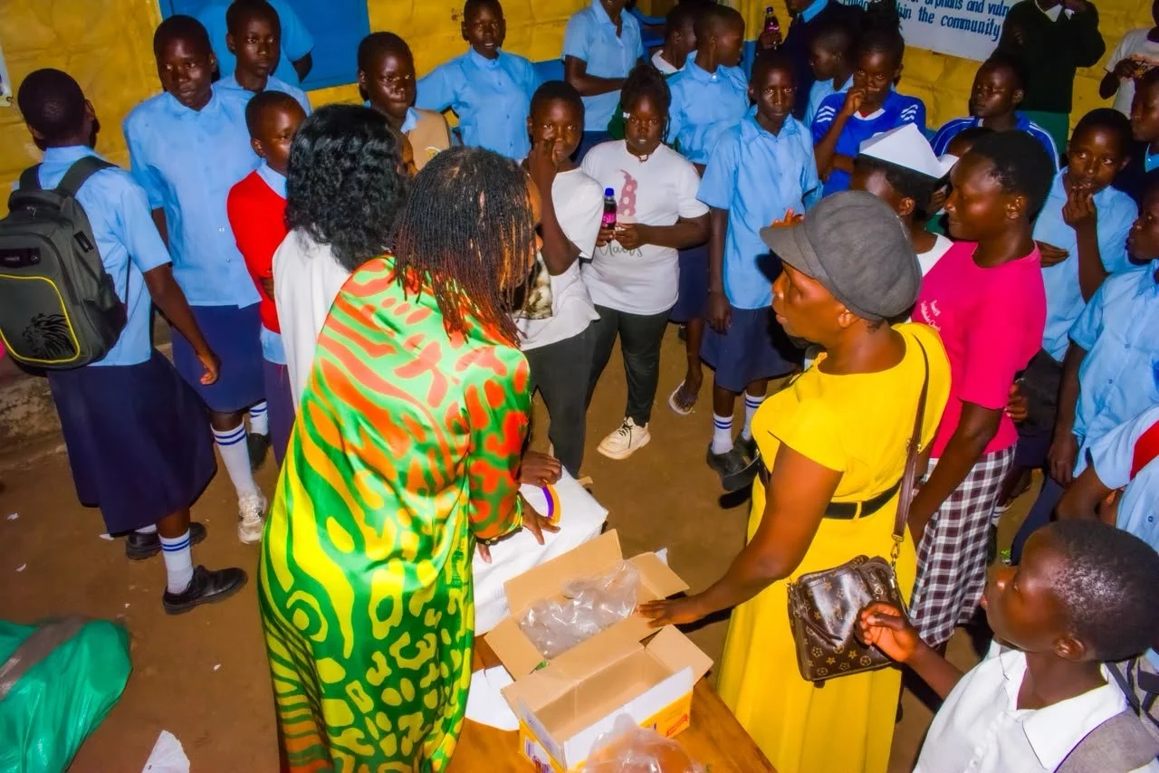 Group of children and two women gathered around a table in a classroom. The women appear to be distributing items from boxes to the children, who are wearing school uniforms. The classroom has a yellow wall and a message about community outreach is visible in the background.