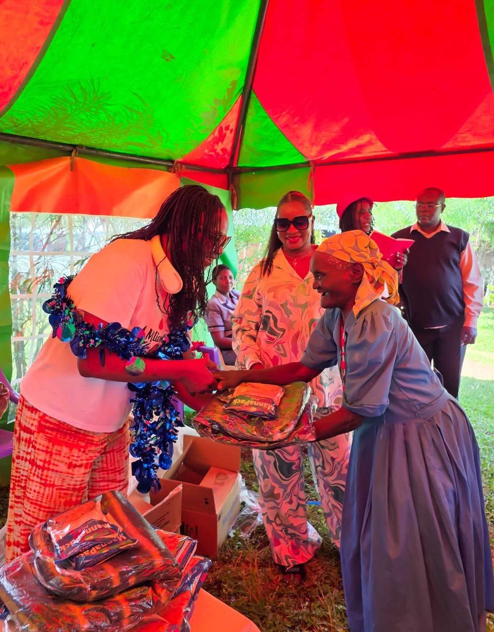 A group of people under a colorful tent, where a woman is handing over a package to a smiling girl dressed in a school uniform. Other adults are standing and sitting nearby.
