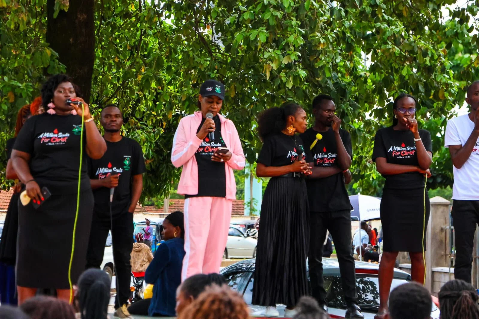 Group of women standing on a platform outdoors, some speaking into microphones, wearing black t-shirts with a religious slogan, with one woman in pink attire, and a large green leafy tree in background, during a public gathering.