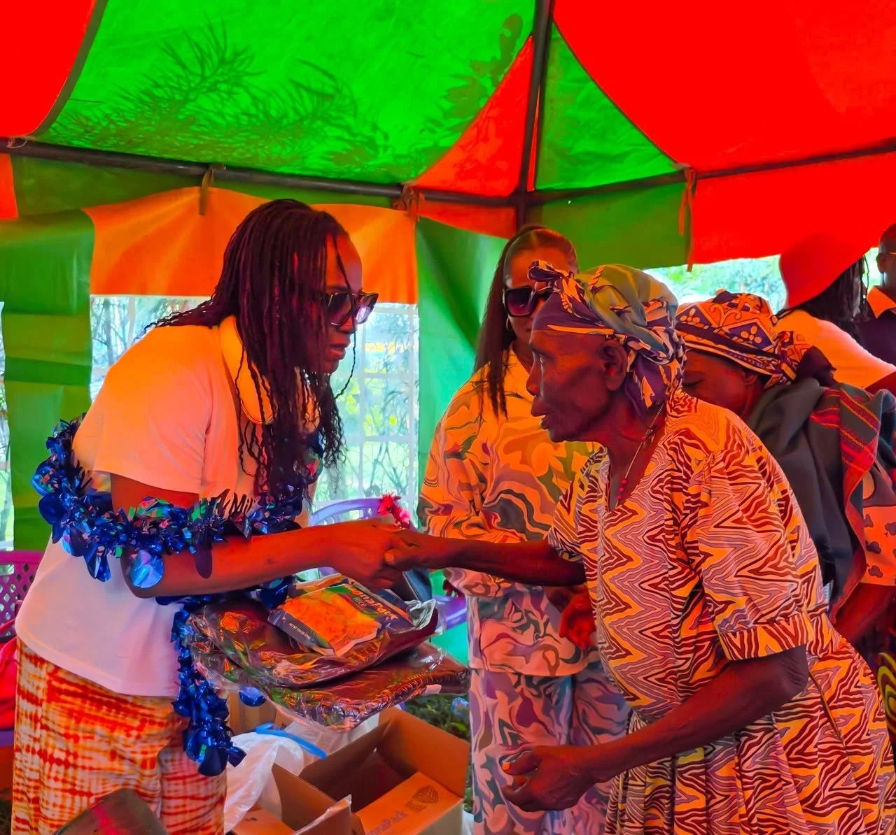A young woman with long braided hair and sunglasses hands a wrapped gift to an elderly woman wearing a patterned headscarf and dress under a colorful tent during an outdoor event.
