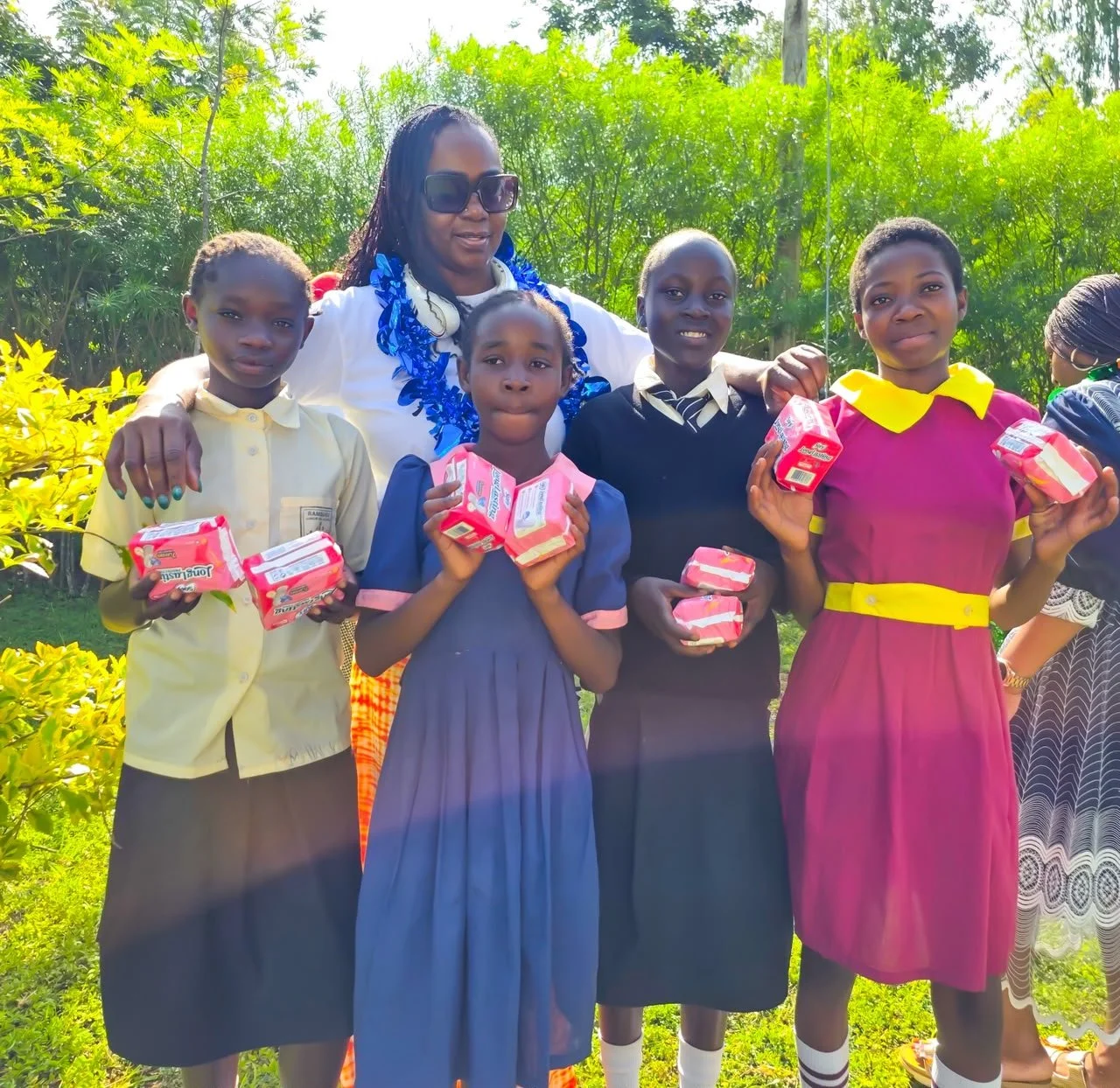 A group of five children and an adult woman standing outdoors, holding small packages of milk, with sunny weather and green foliage in the background.