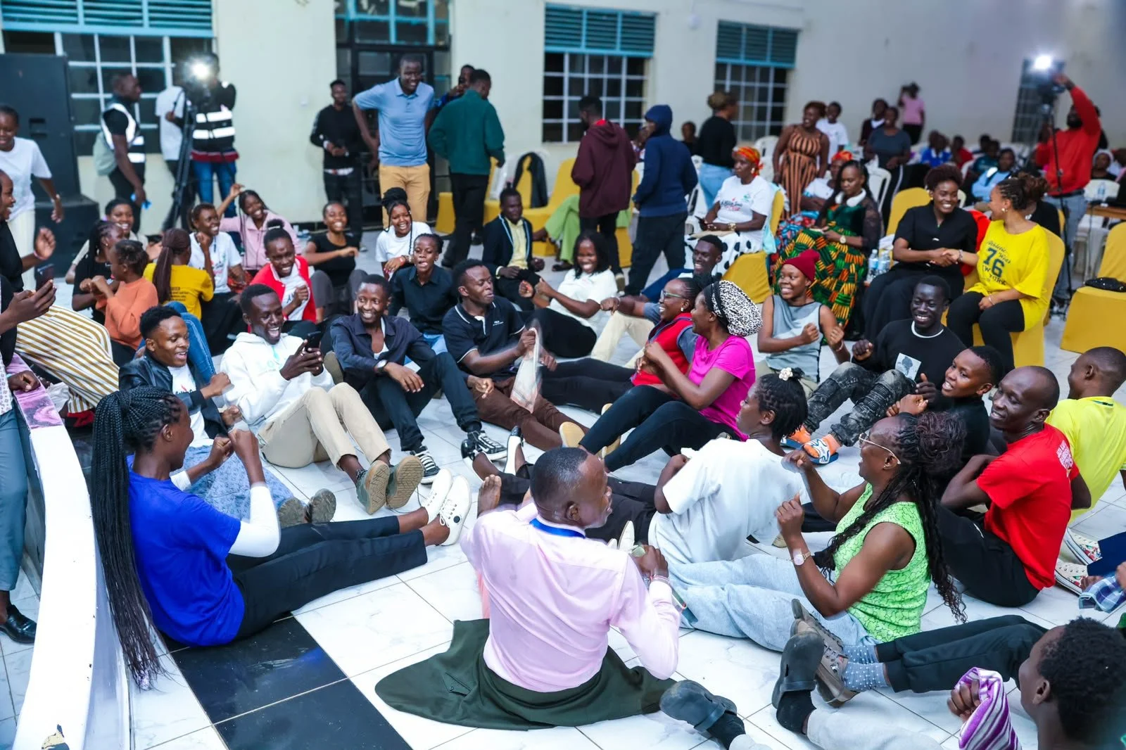 A large group of people sitting on the floor and in chairs, smiling and laughing during a social gathering or event inside a room with large windows.