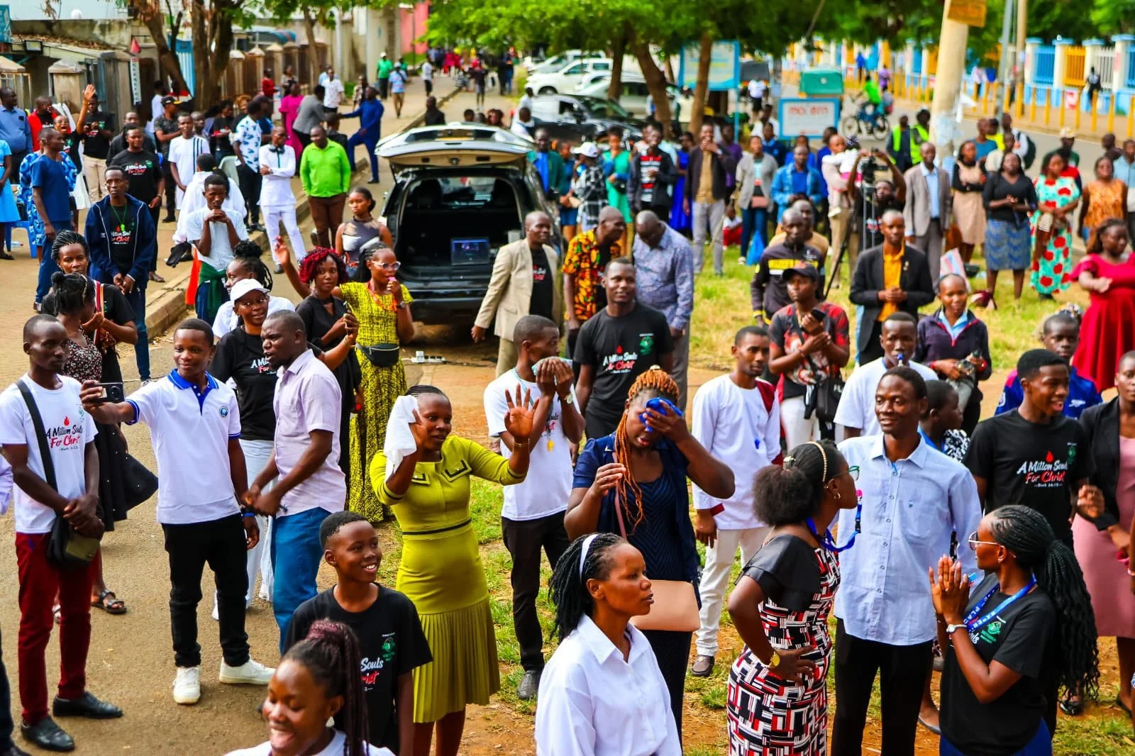 A large crowd of people gathered outdoors in a park or street, with many smiling and engaging with each other. Some are taking photos, and a few are waving or covering their faces. There are trees, parked cars, and colorful buildings in the backgroun