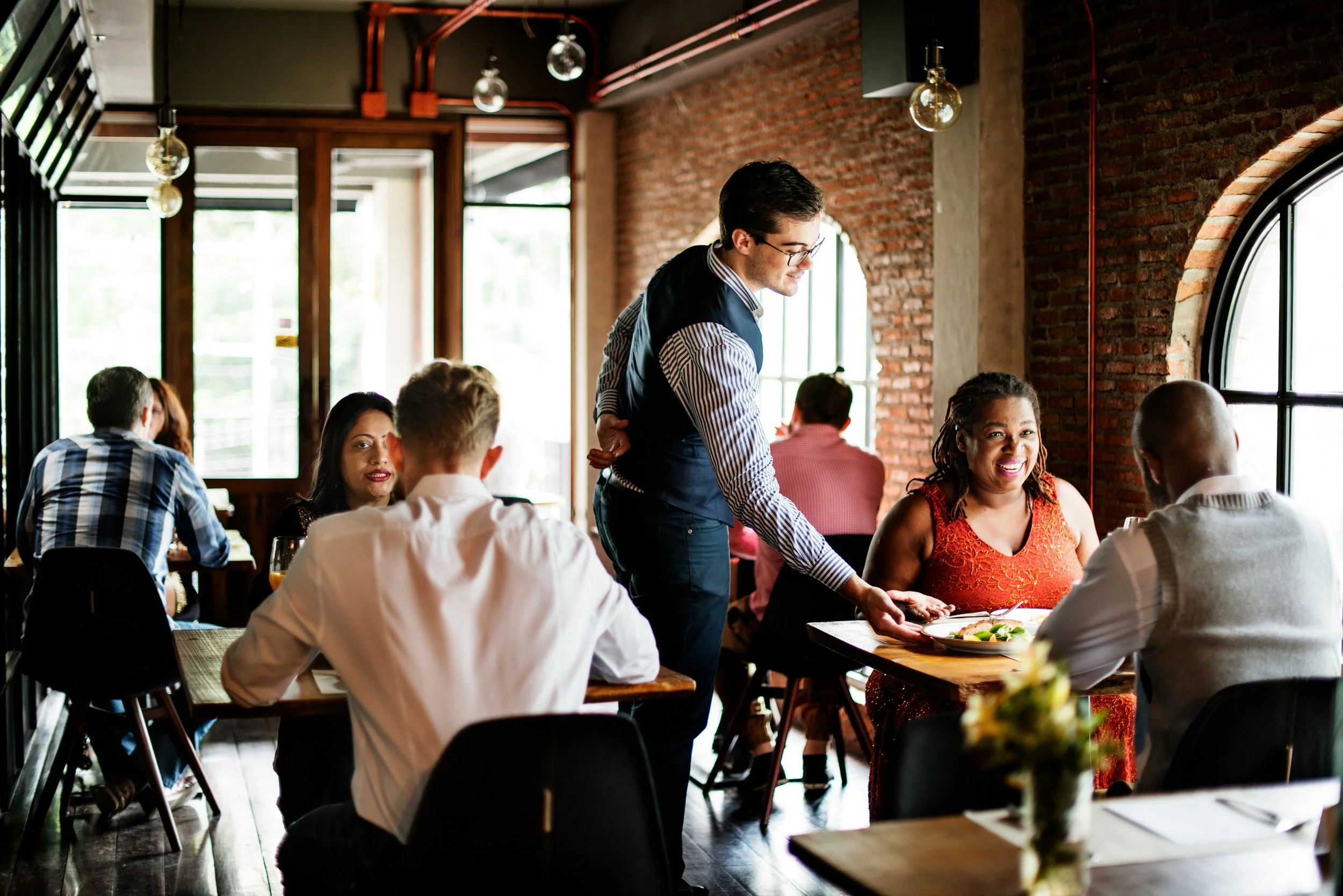 A group of diverse people in a restaurant enjoying a meal, with a waiter serving food. The restaurant has brick walls, large windows, and hanging light fixtures.
